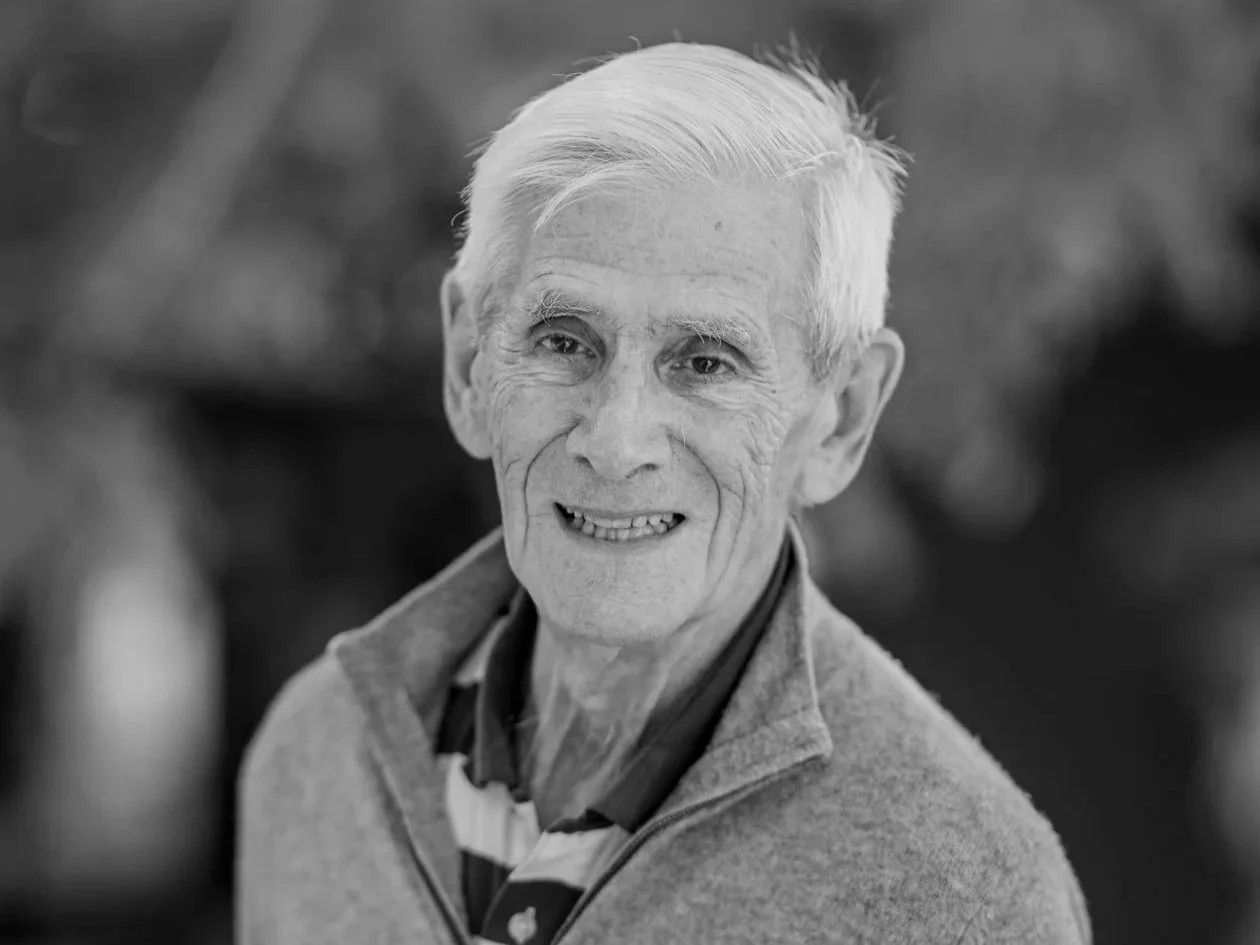 Black and white photo of man with short silver crewcut hair, smiling at camera, wearing stripped polo shirt underneath grey zip front jacket