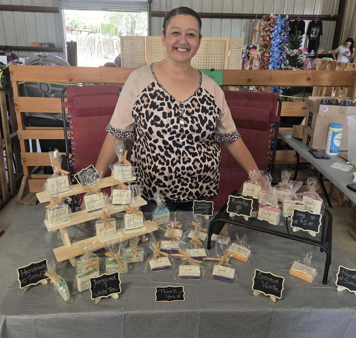 A woman smiling at a craft fair booth displaying handmade soaps with decorative labels and small chalkboard signs indicating scents and prices, set up indoors with a table covered in a gray tablecloth.