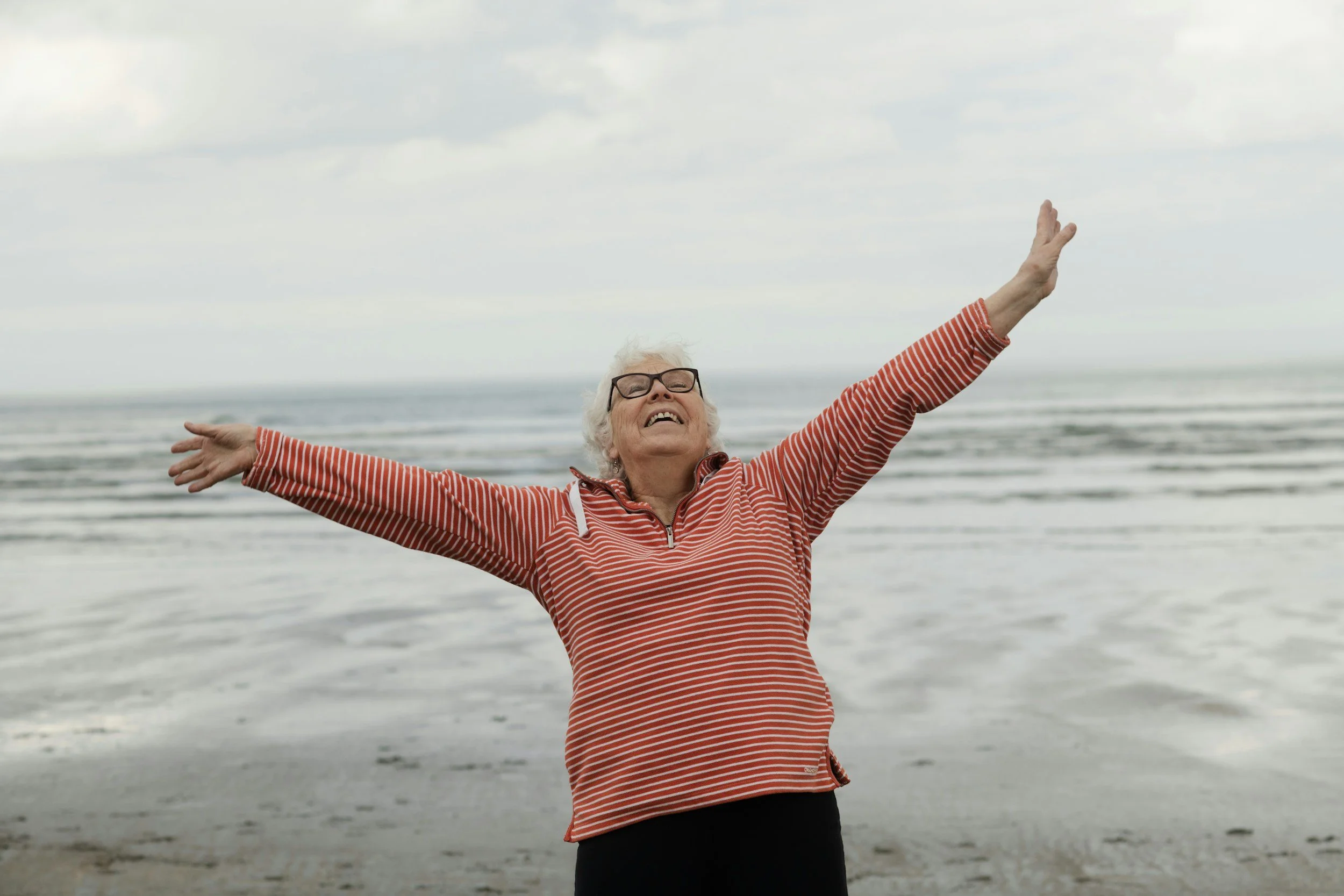 An elderly woman with white hair, glasses, and a red and white striped jacket standing on a beach with arms outstretched and smiling.