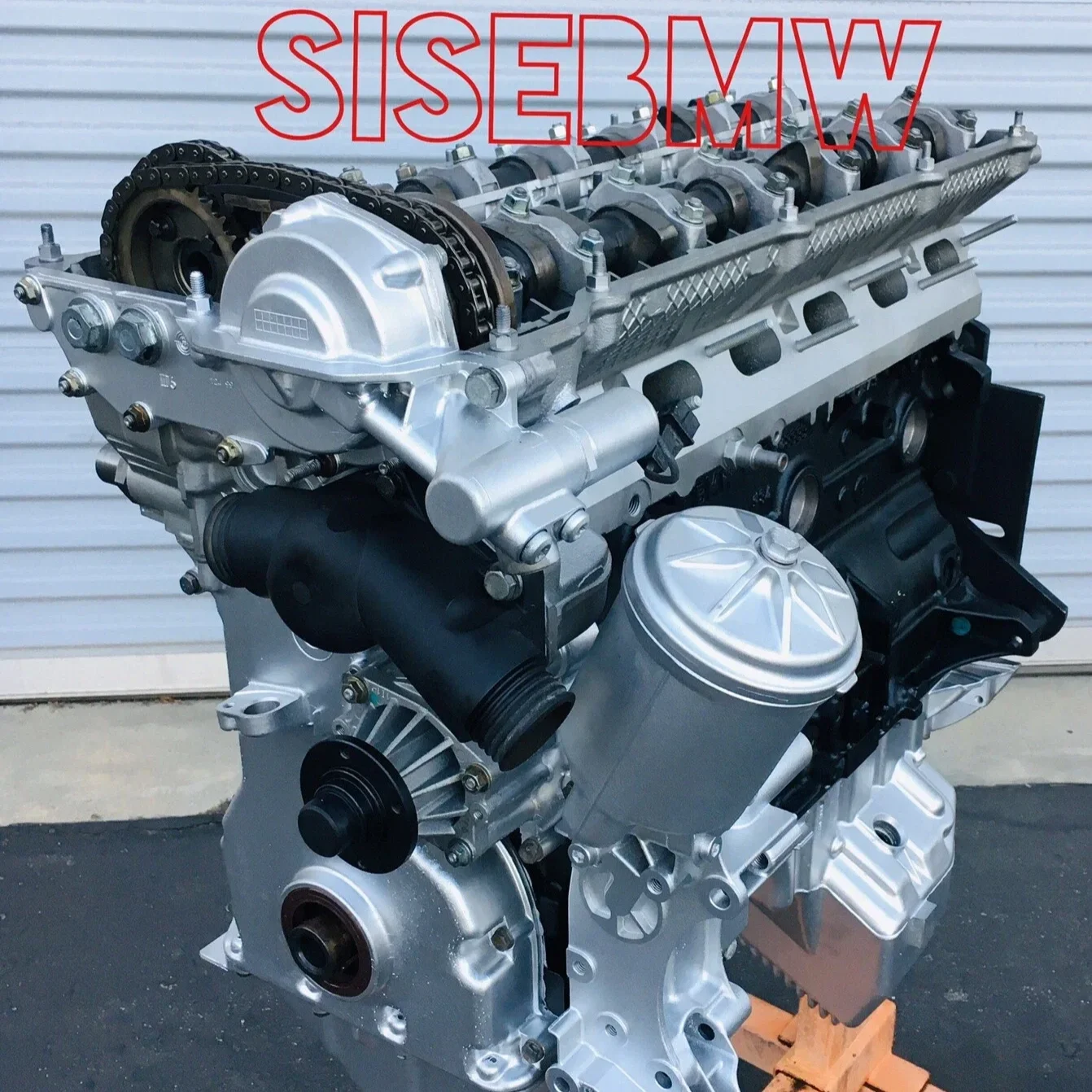 A new BMW engine with the text SI5EBMW overlayed in red, placed in front of a white garage door.