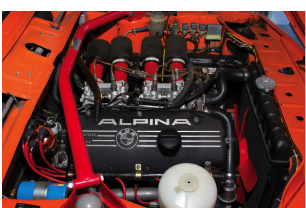 Close-up of a car engine with a black Alpina cover and red hoses, surrounded by engine components.