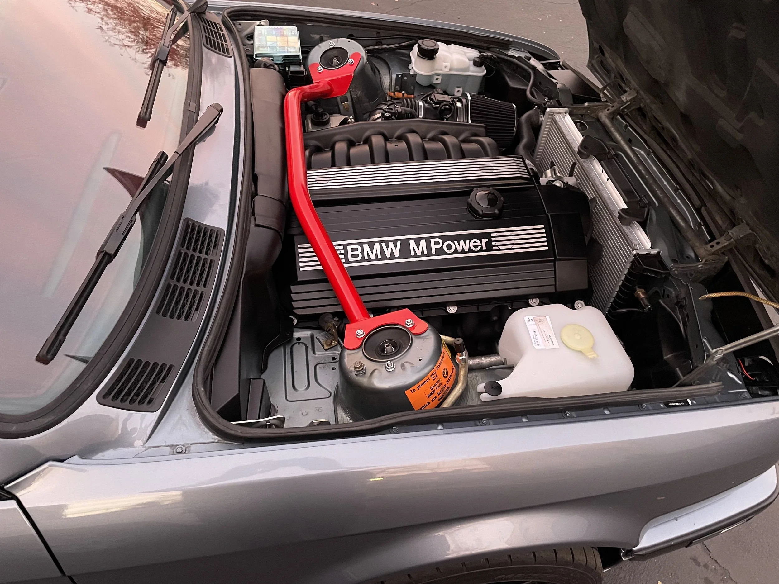 A car engine bay with a black BMW M Power engine cover, a red strut brace, and various engine components, including a coolant reservoir and an intercooler.