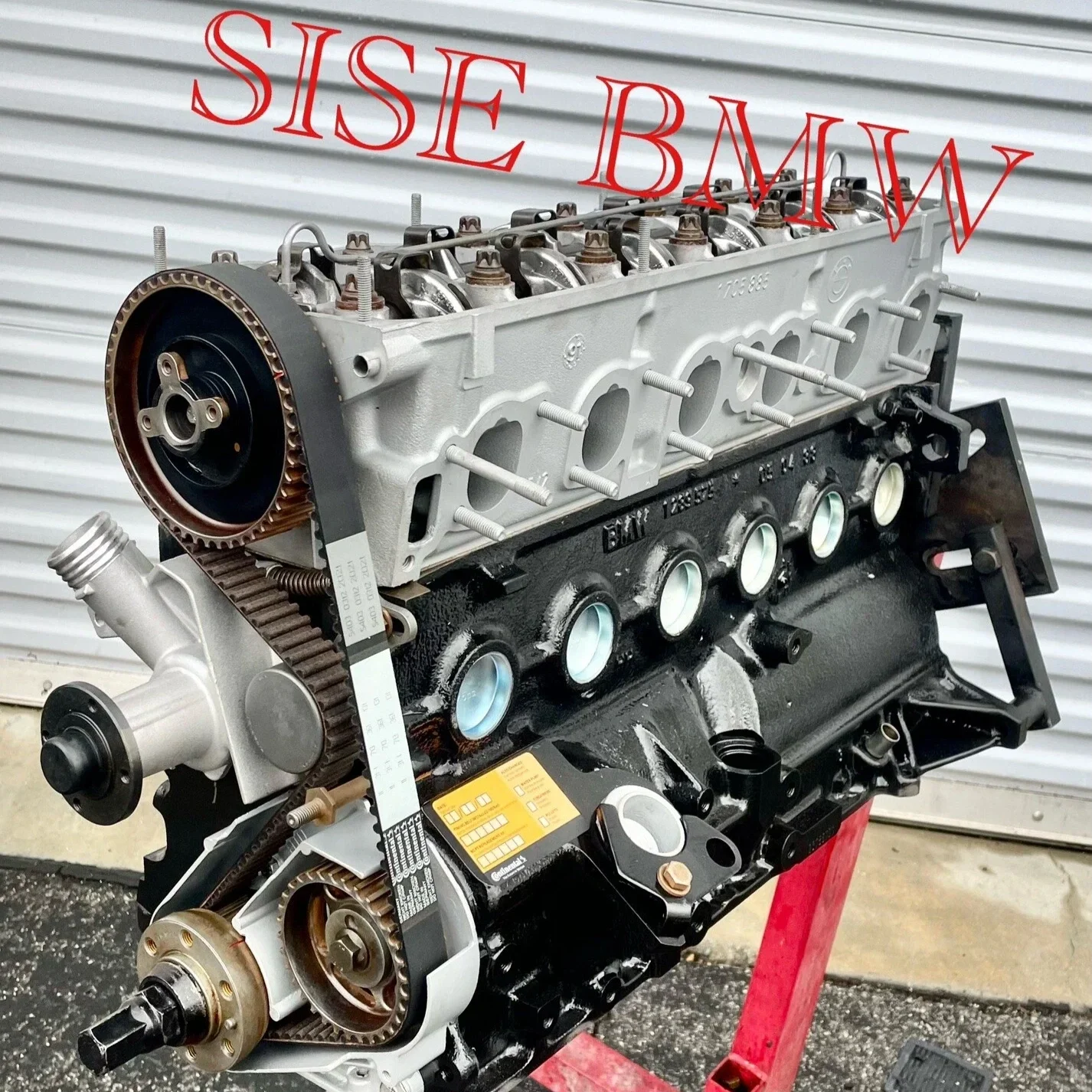 An engine block, transmission, and timing belt on a stand outside, with a corrugated metal wall in the background.