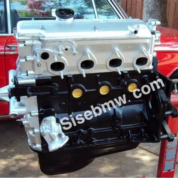 A car engine with a silver top and black bottom, displayed outdoors with a red vehicle and trees in the background.