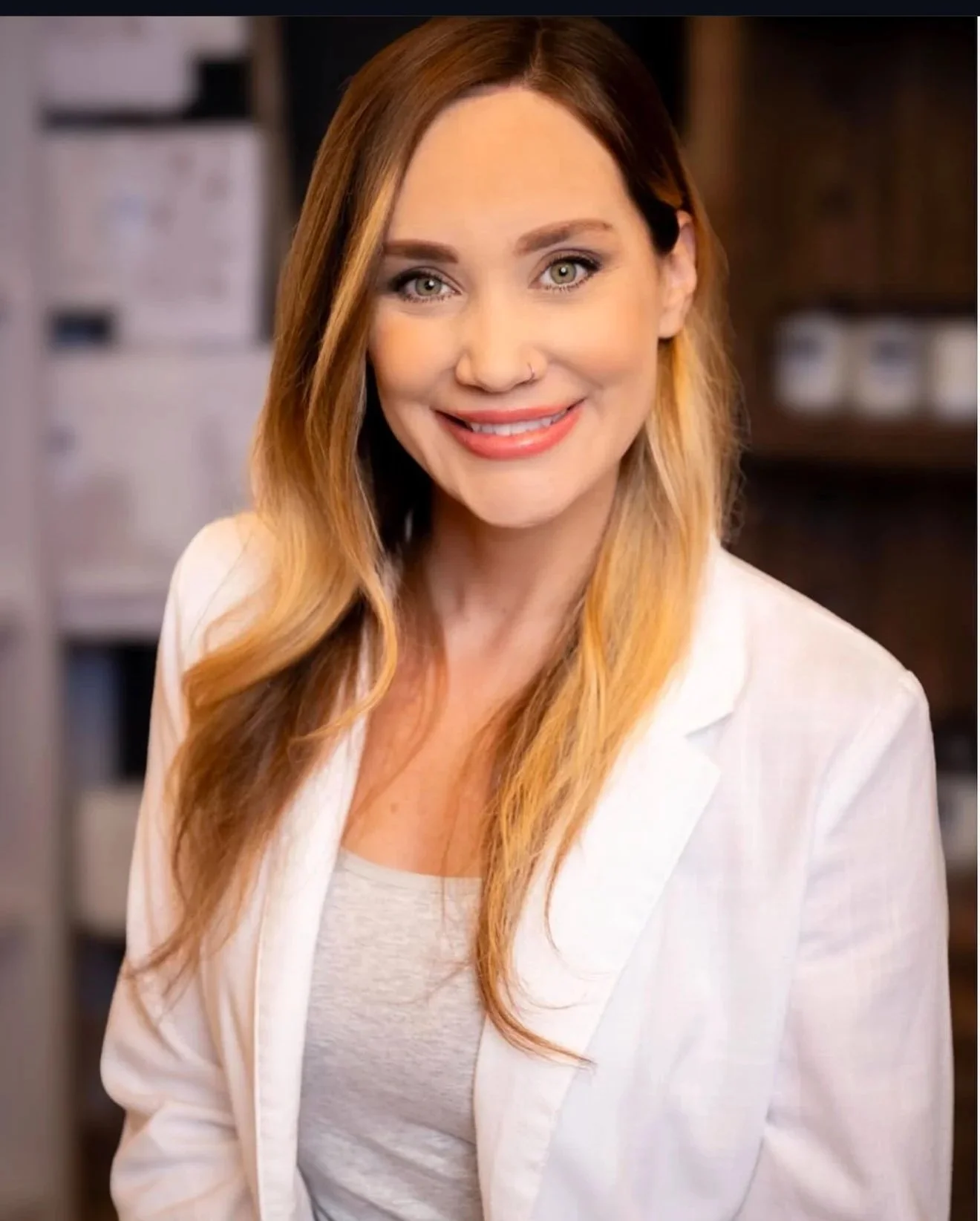 A woman with long, wavy, reddish-brown hair and light skin, smiling and wearing a white blazer over a light gray shirt, standing indoors with a blurred background.