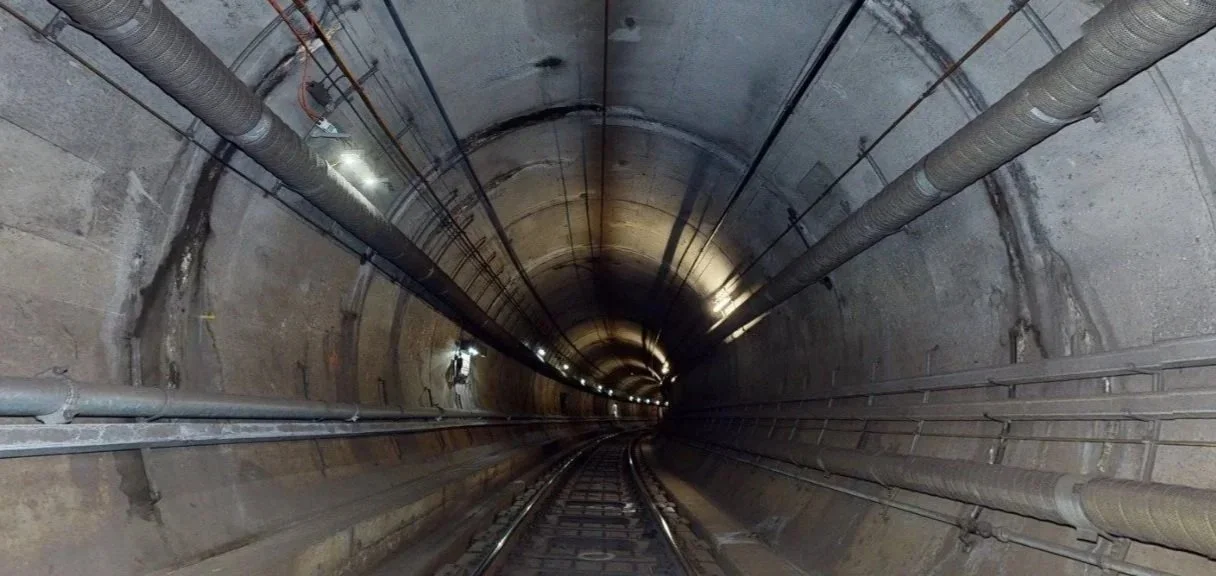 Looking down a long underground tunnel with train tracks and pipes along the walls and ceiling.