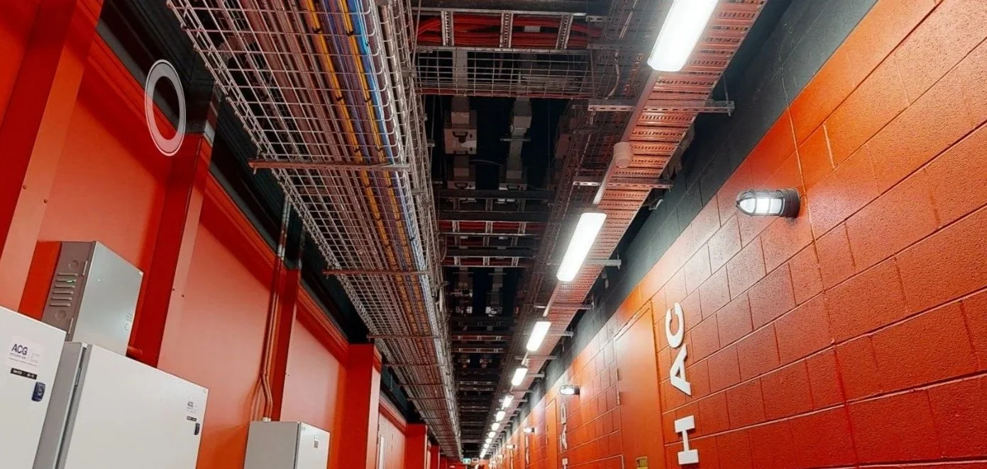 Industrial hallway with orange brick walls, ceiling mounted electrical wiring and metal cable trays, white electrical cabinets, and exterior lighting.