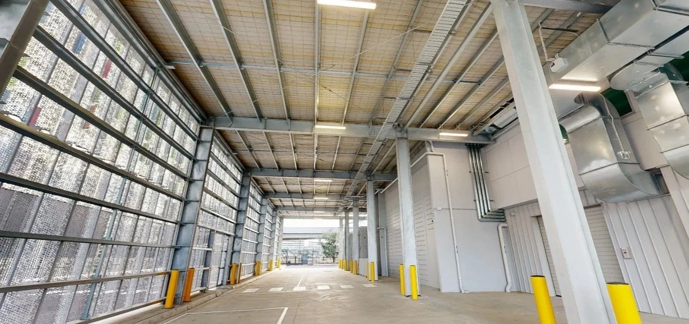 An empty loading dock area with high walls and overhead metal roof, yellow safety guards along the edges, and a small glimpse of the outside with trees and a cloudy sky.