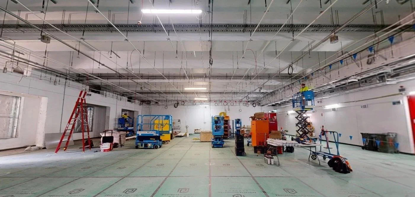 Construction workers installing ceiling fixtures in an unfinished indoor space with construction equipment, lifts, and scaffolding.