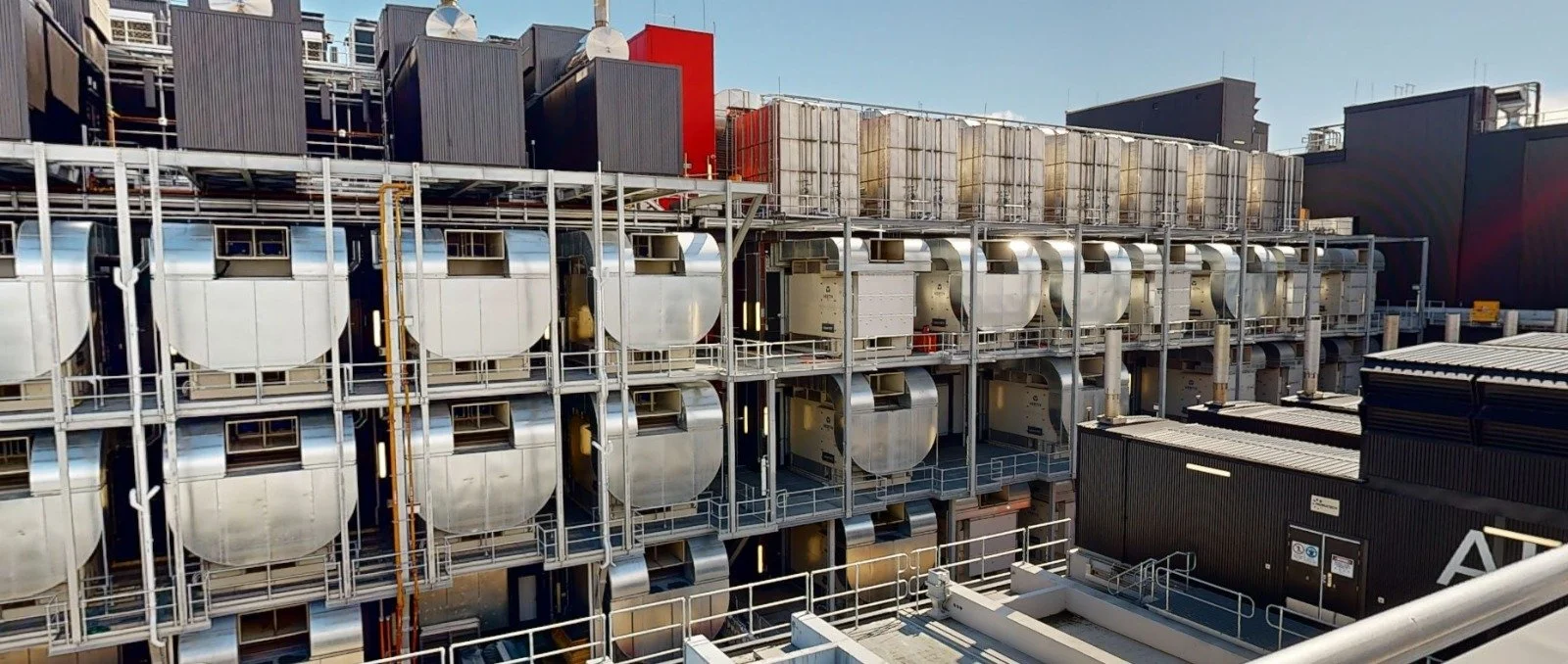 Industrial rooftop with large metallic ventilation units, pipelines, and structural framing, set against a blue sky.