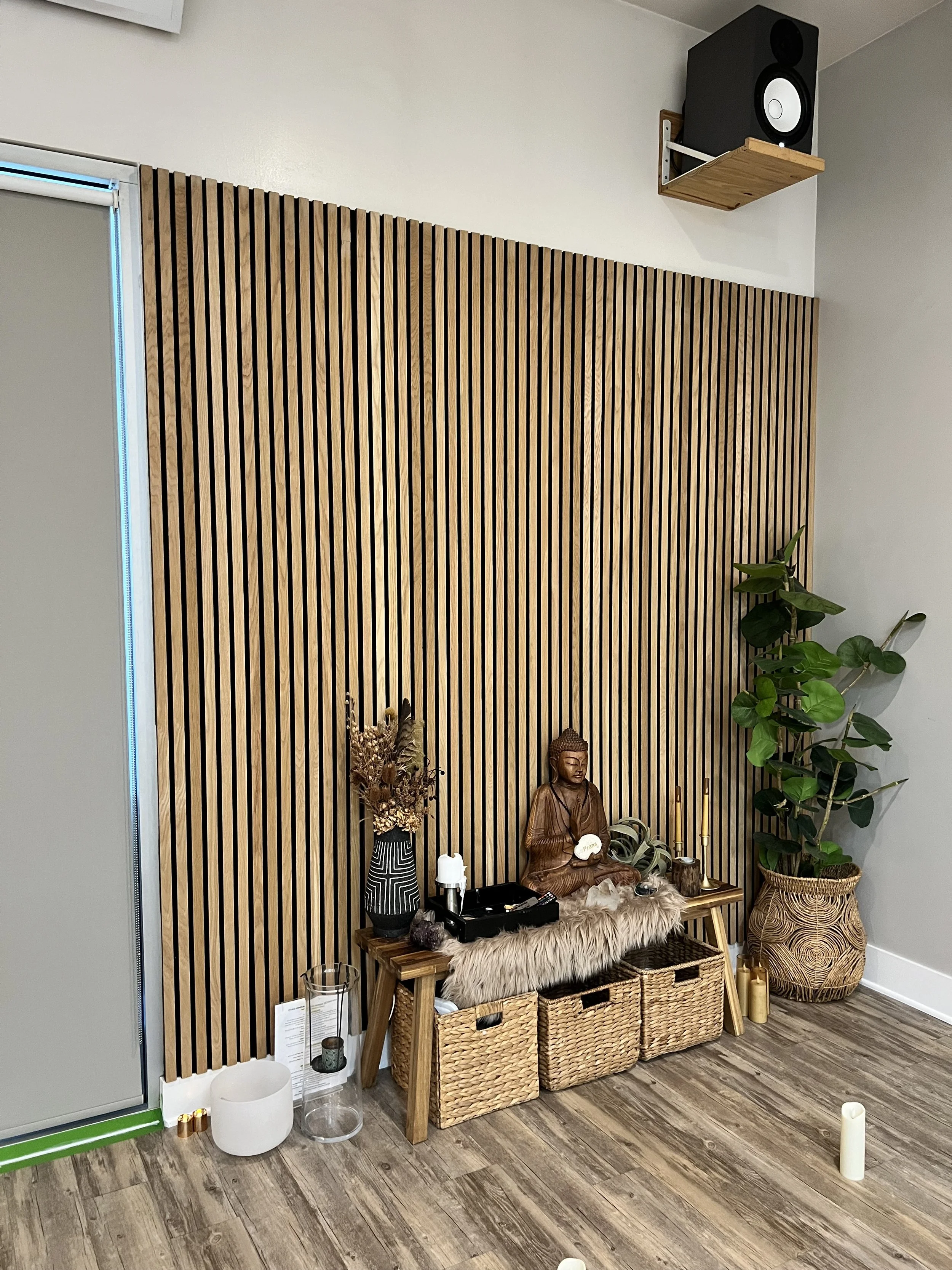 A yoga or meditation space with a wooden slat wall, a small altar with a Buddha statue, candles, plants, and woven baskets. There are decorative objects and a speaker mounted high on the wall.