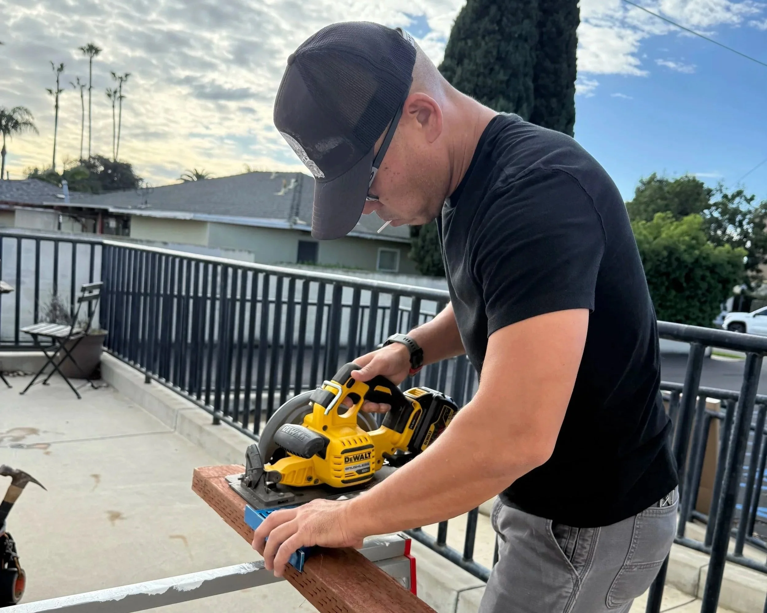 A man wearing a black t-shirt and gray jeans using a yellow DEWALT power tool on a piece of wood on an outdoor balcony.