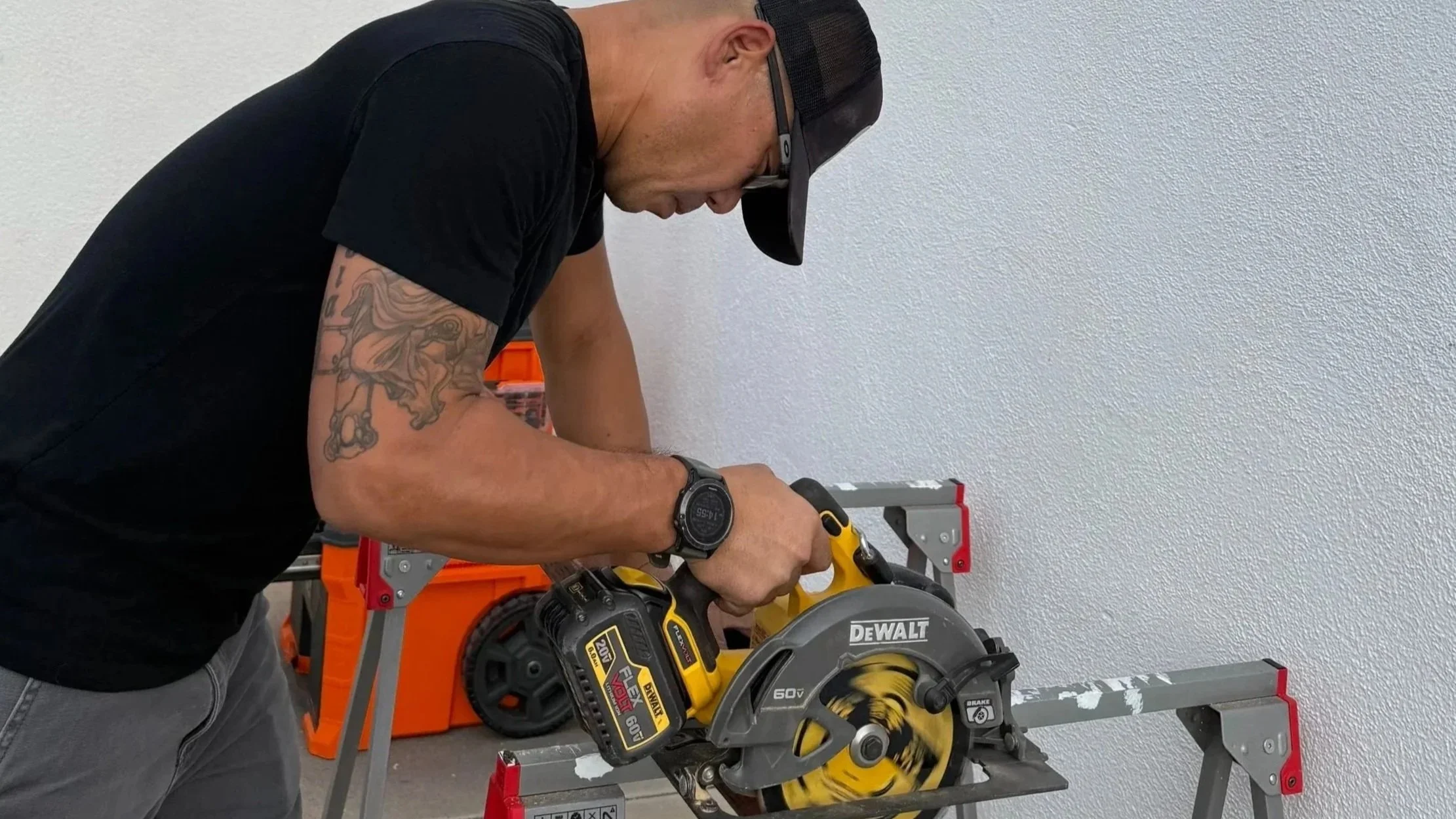 A man in a black shirt using a yellow DeWalt cordless circular saw to cut a piece of wood or metal on a worktable. The man is wearing a black baseball cap and a black wristwatch. There are orange and gray toolboxes in the background.