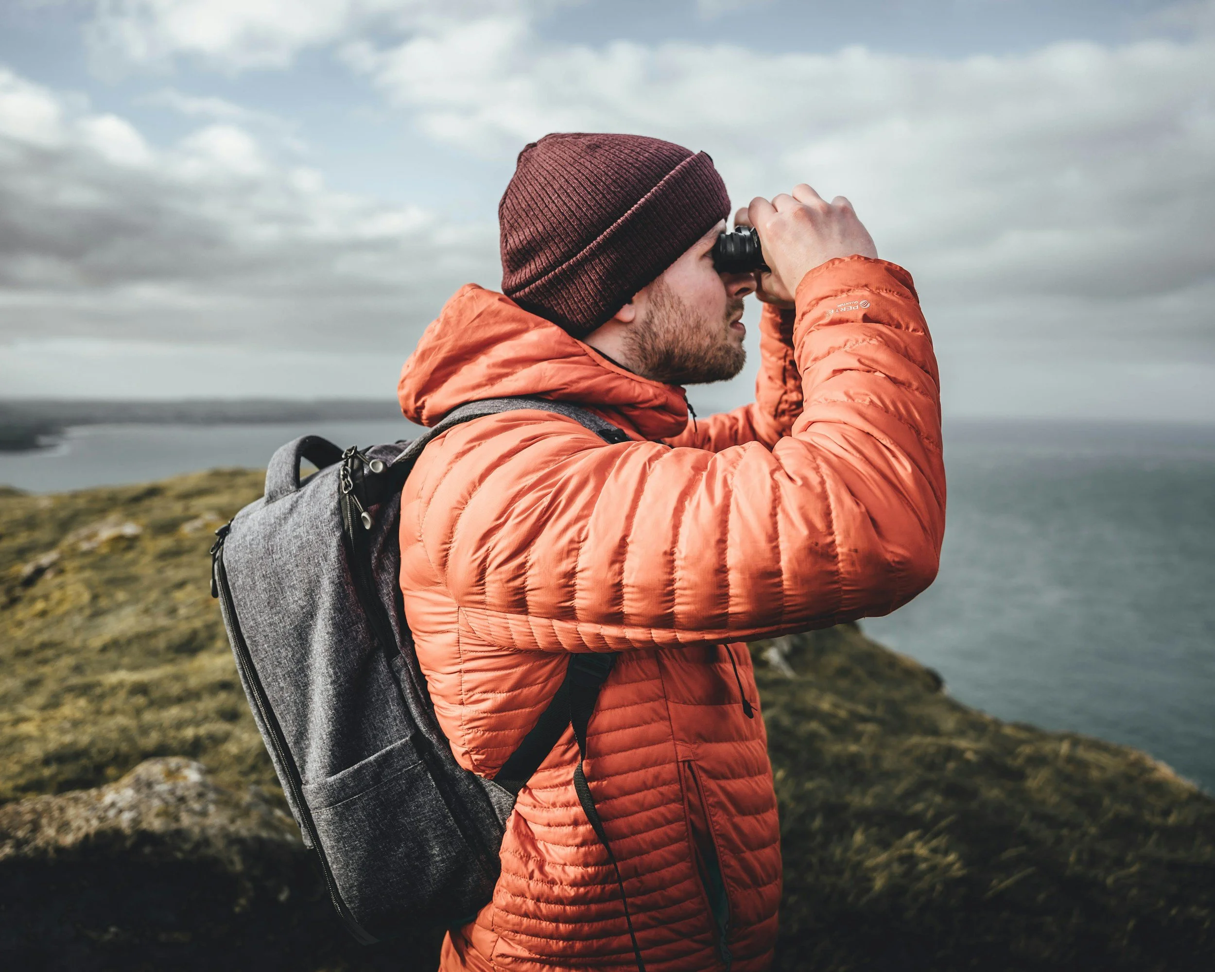 A man wearing an orange puffer jacket and a maroon beanie using binoculars outdoors on a hillside during daytime, with cloudy skies and a body of water in the background.