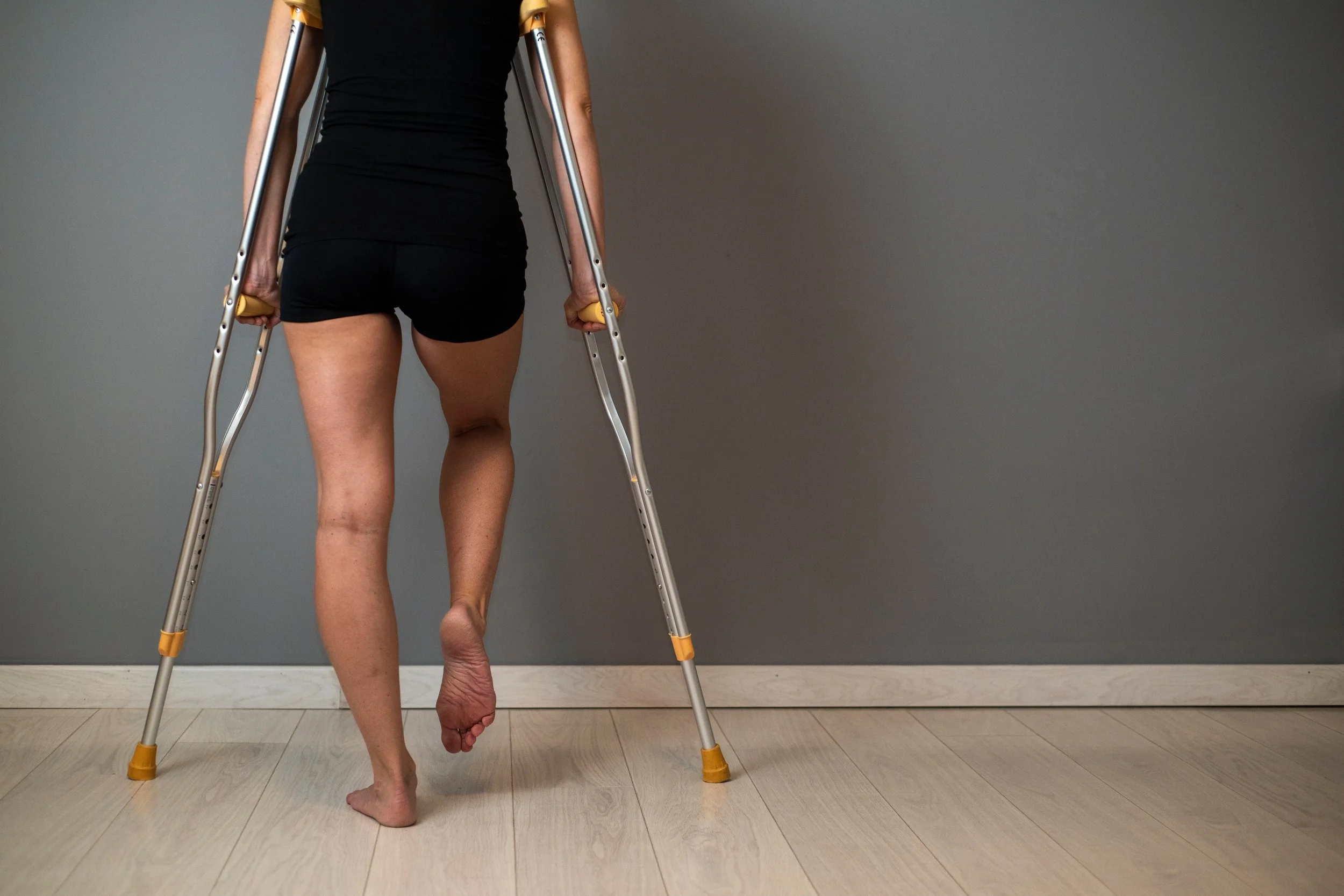A person using crutches, wearing a black tank top and shorts, walking indoors with gray wall background and light-colored wooden floor.