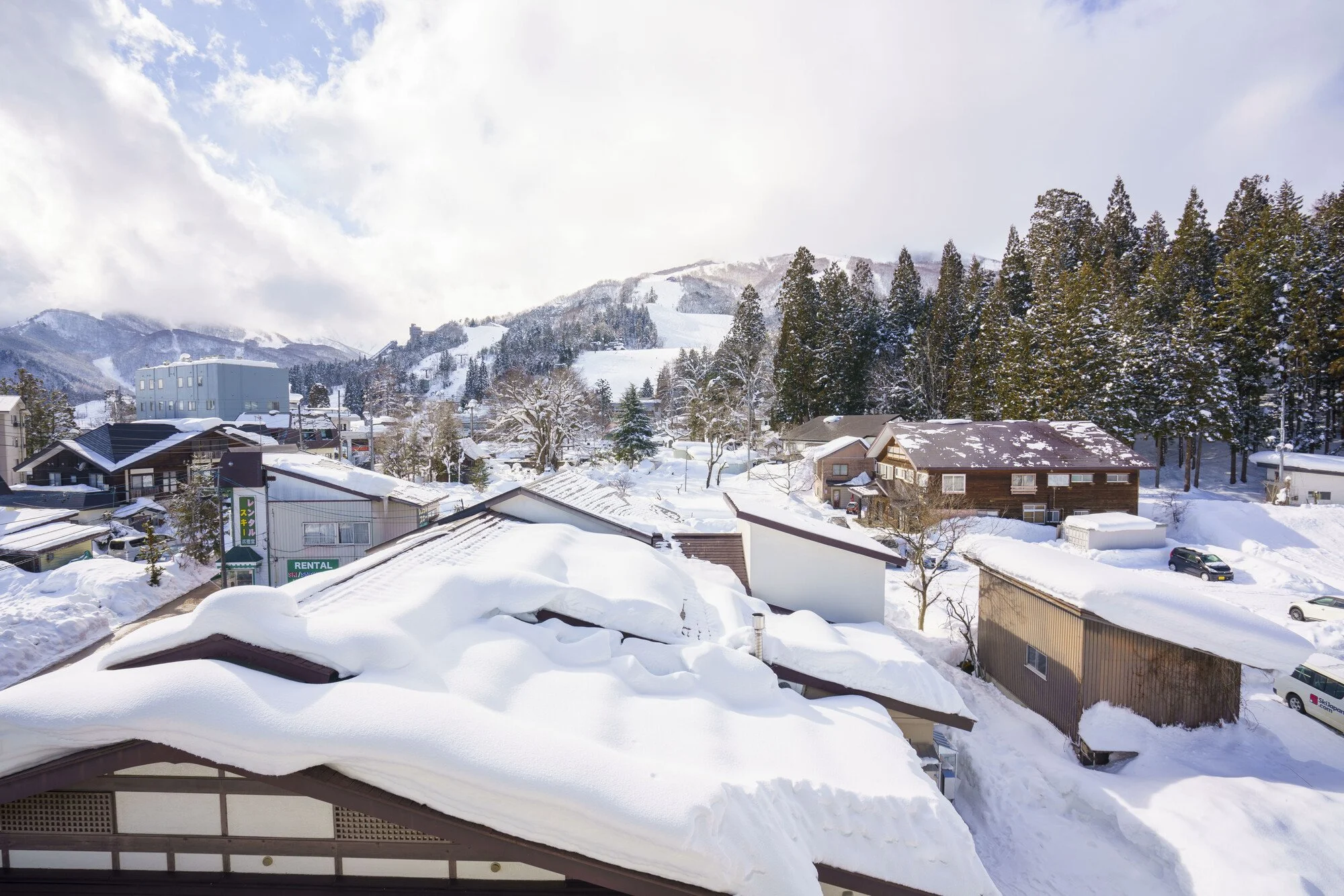 Snow-covered rooftops in a mountain town with trees, houses, and cars, under a partly cloudy sky.