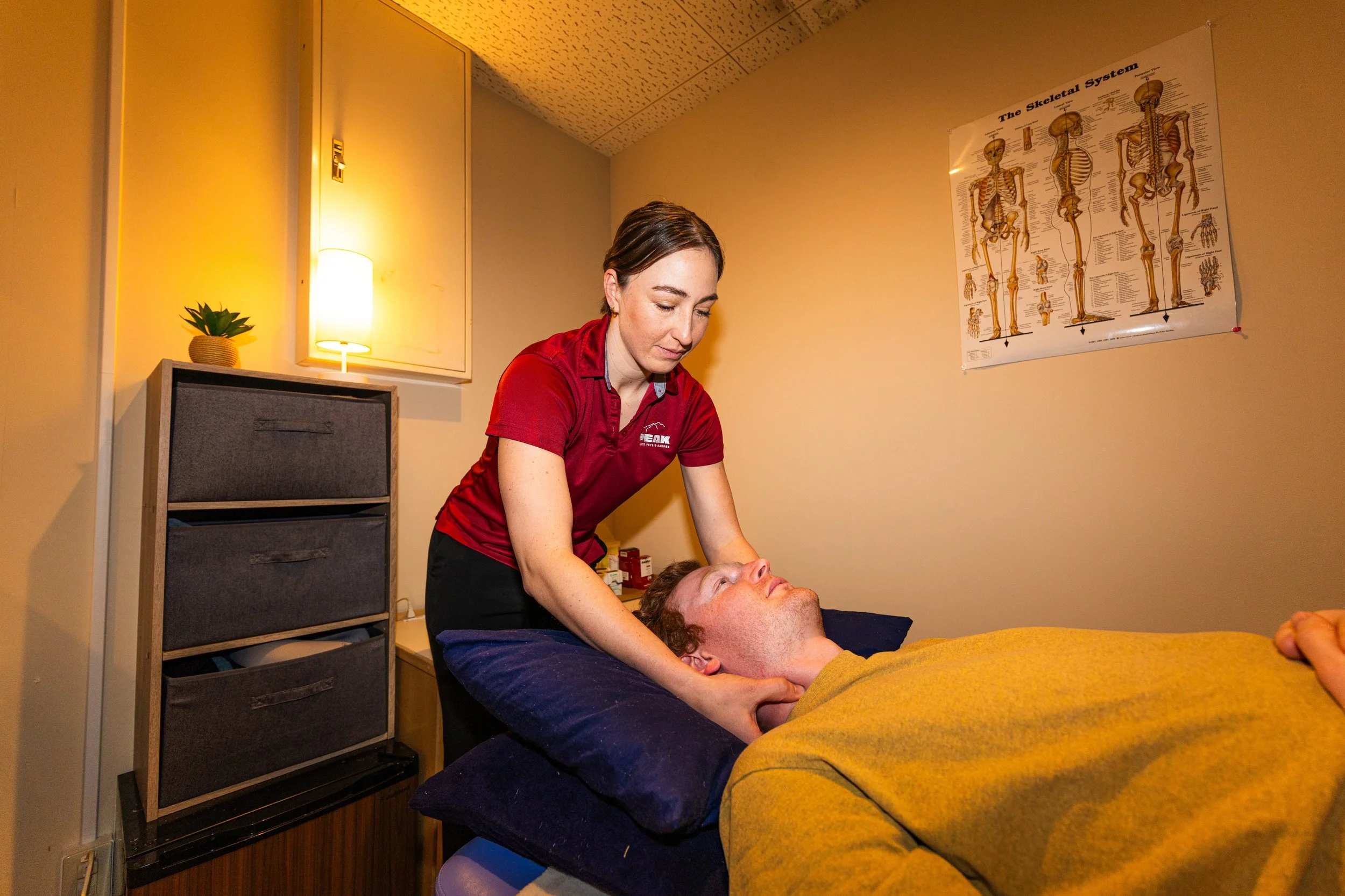 A woman in a red shirt providing a massage to a man lying on a massage table covered with a yellow blanket, in a room with a skeleton anatomy chart on the wall.