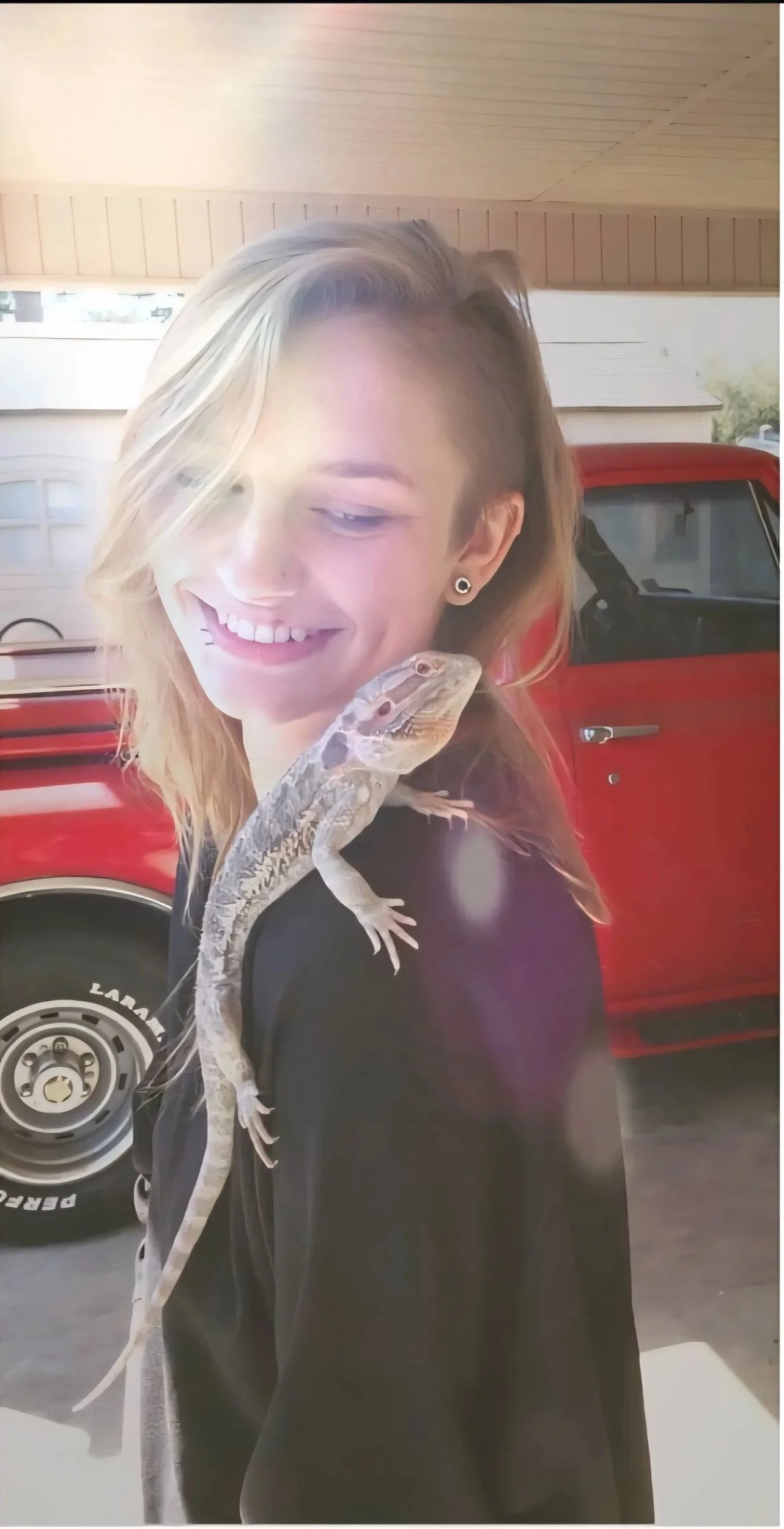 A person with blonde hair smiling and looking down, standing outside near a red vehicle, with a gecko lizard on their shoulder.