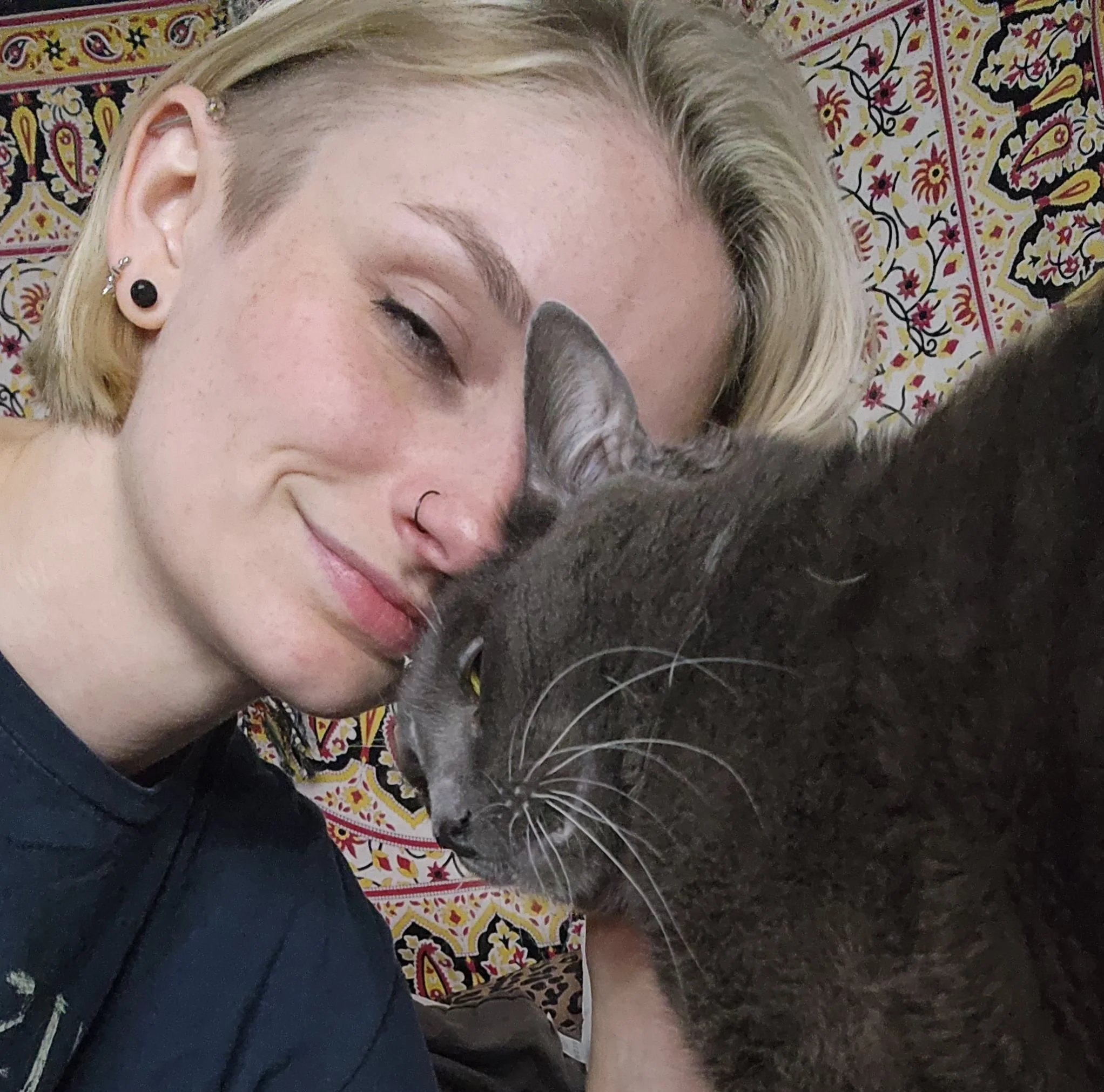 A woman with blonde hair and earrings leaning close to a gray cat on a patterned fabric surface.