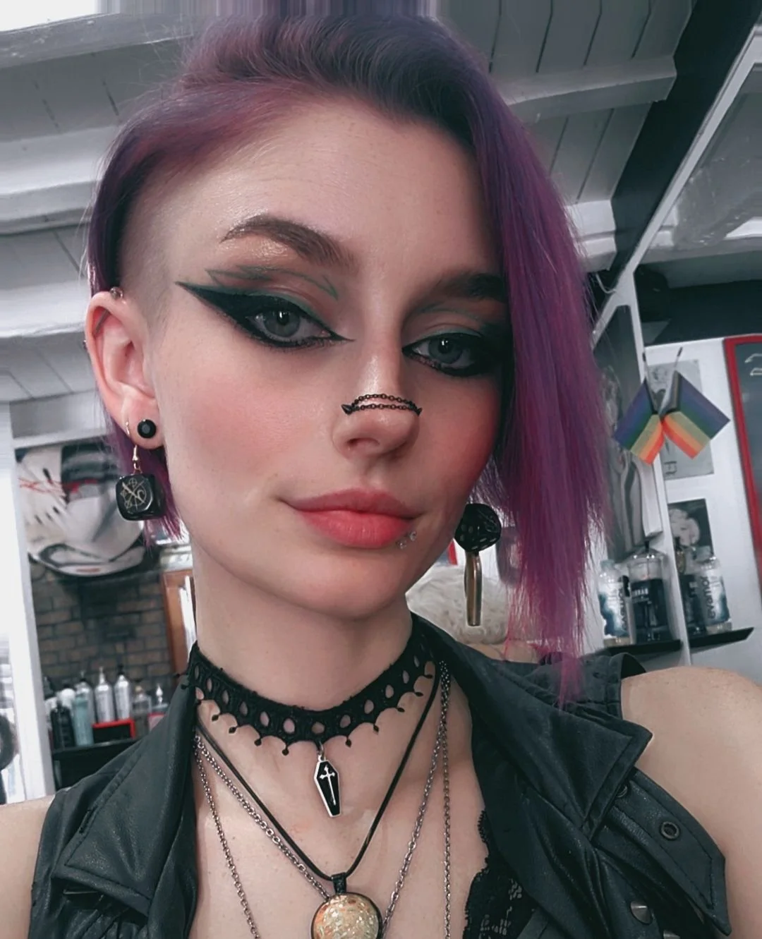 A young woman with purple hair and makeup, wearing gothic jewelry, with rainbow flags and hair products in the background.
