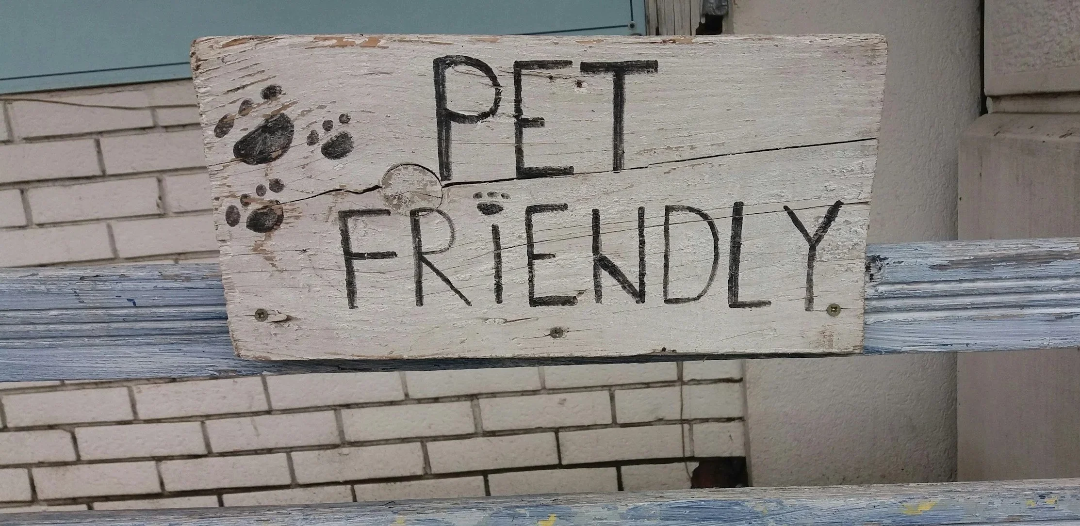 A rustic wooden sign painted white with black handwritten text that reads 'PET FRIENDLY'. There are paw print illustrations to the left of the text. The sign is mounted on a distressed white wooden beam, with a brick wall in the background.