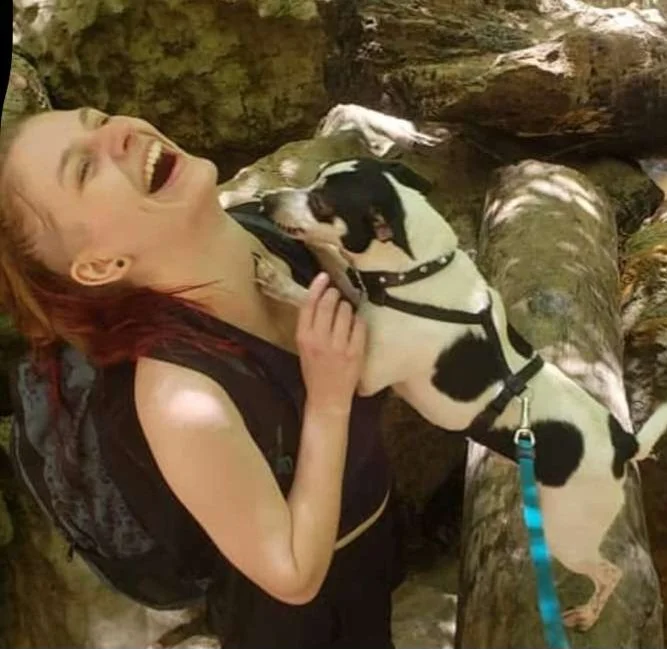A woman with red hair laughing and smiling while hugging a small black and white dog, with rocks and a log nearby.