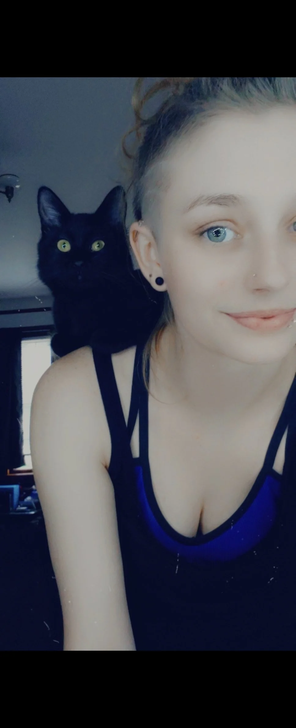 A young woman with blonde hair, blue eyes, and a black tank top with blue accents, taking a selfie with a black cat sitting on her shoulder, indoors in front of a window with blinds.