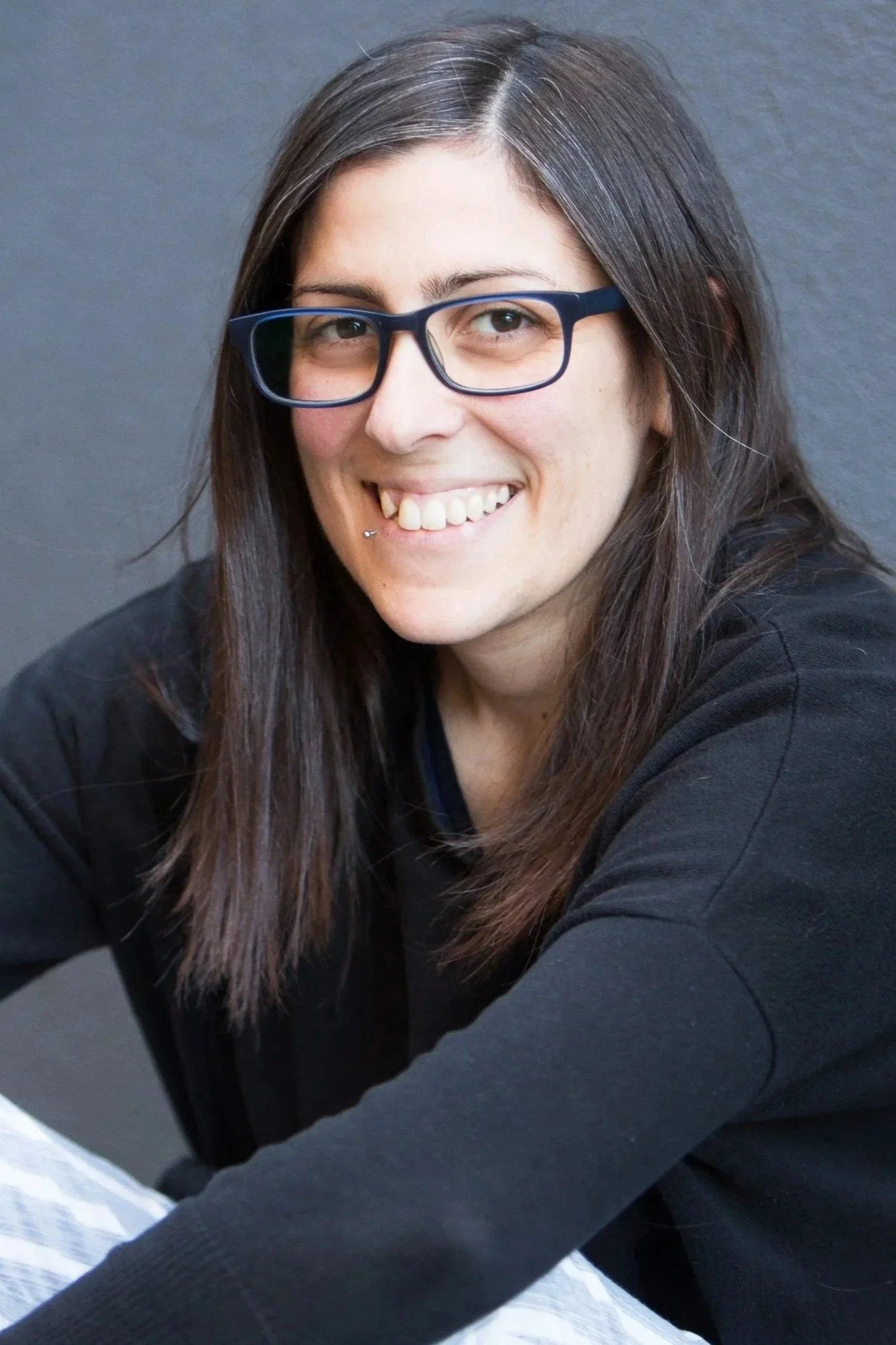 A woman with long dark hair, glasses, and a piercing smiling at the camera against a plain gray background.