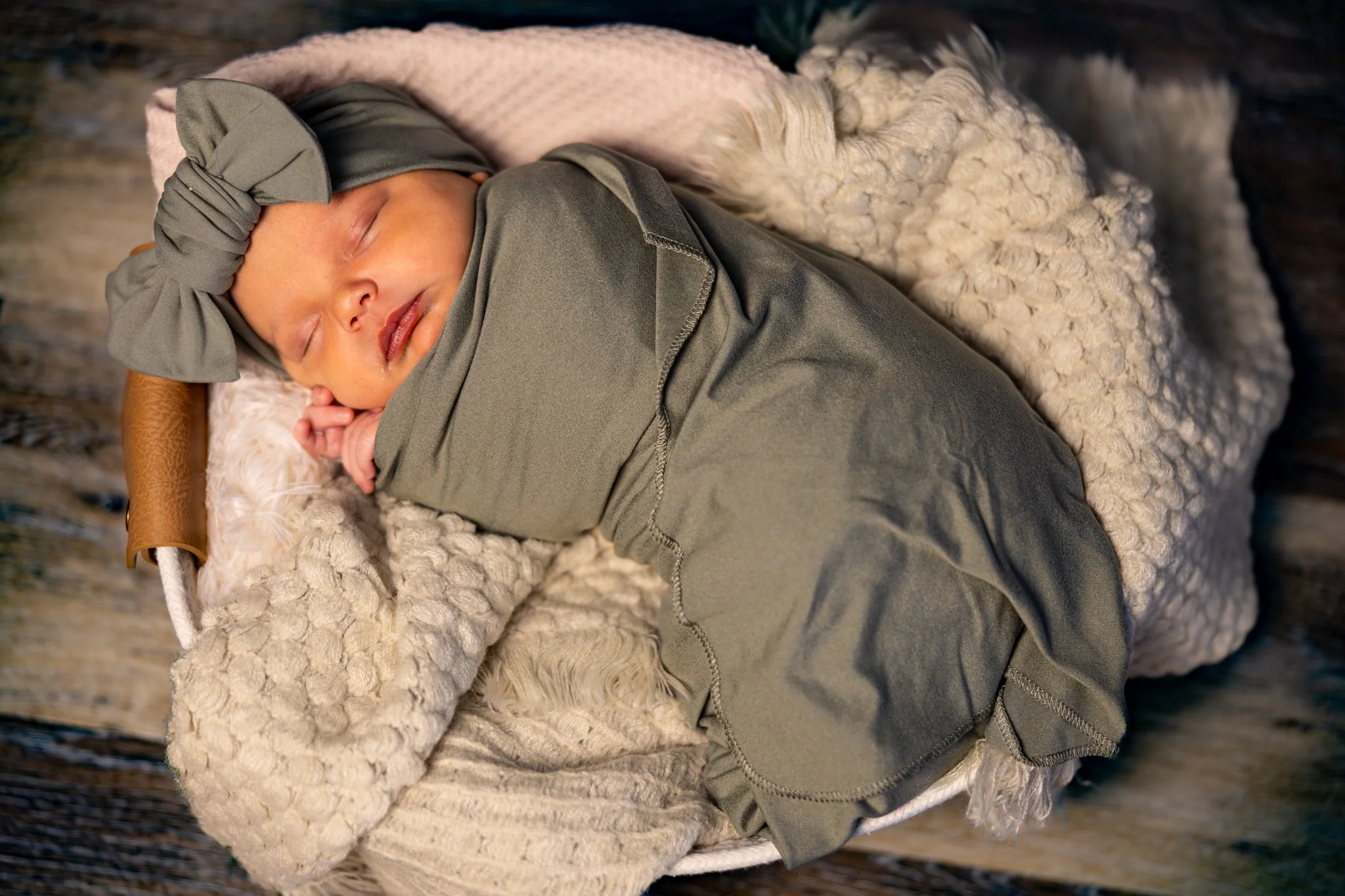 A sleeping baby wrapped in a gray blanket and wearing a gray headband with a bow, lying on a soft, textured cream blanket and a wooden surface background.