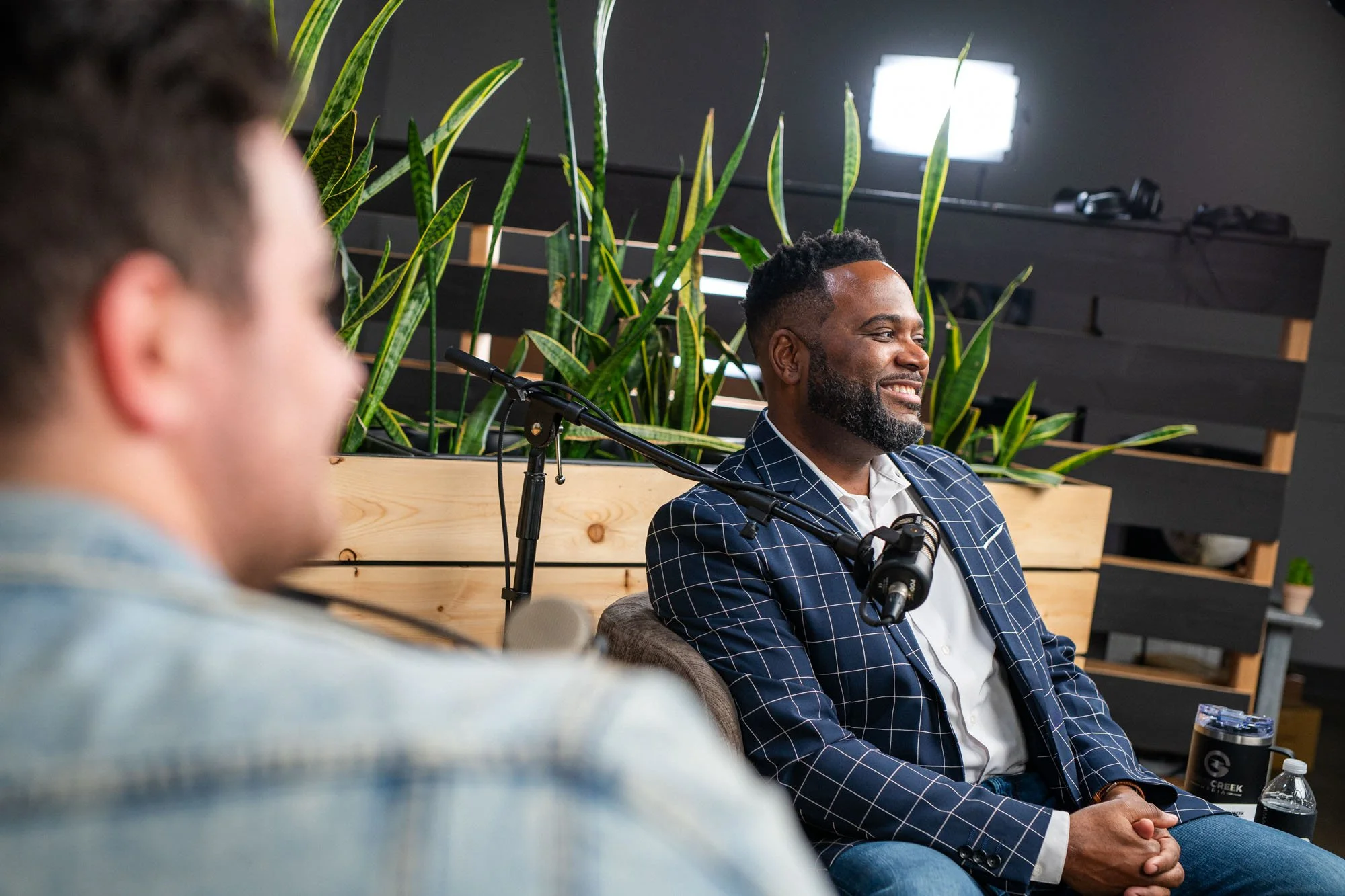 Man in a blue checkered suit smiling during a discussion in a meeting room with indoor plants and a black wall with shelves in the background.