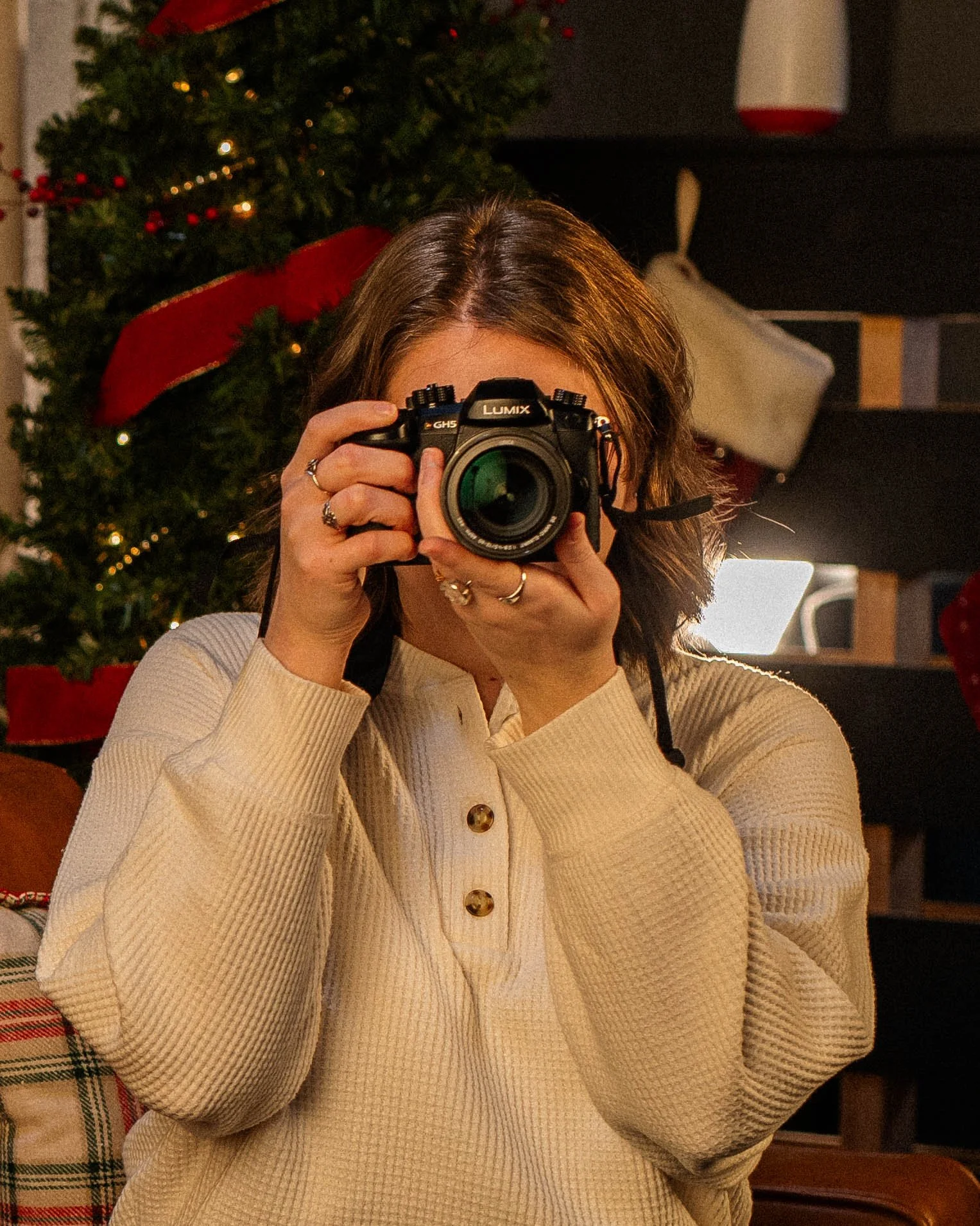 A woman with brown hair taking a photo of herself with a camera in front of a Christmas tree decorated with red ribbons and lights.