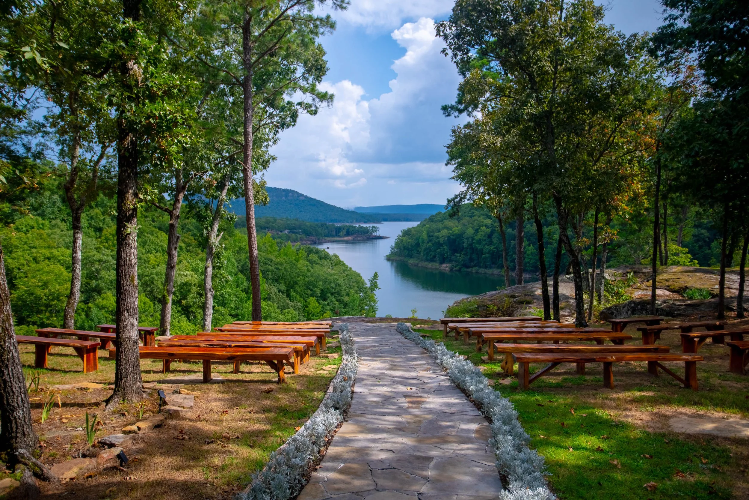 A scenic outdoor view of a river flowing through a lush green forest with a row of wooden benches and a stone pathway in the foreground, tall trees on either side, and a partly cloudy sky overhead.
