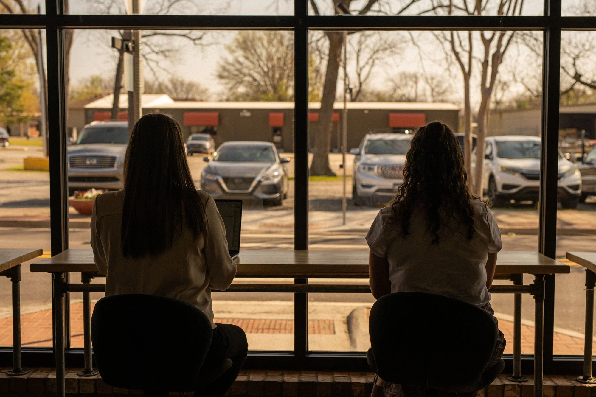 Two women with dark hair sitting at a table facing a large window overlooking a parking lot with several cars and trees, in an indoor cafe or restaurant setting.