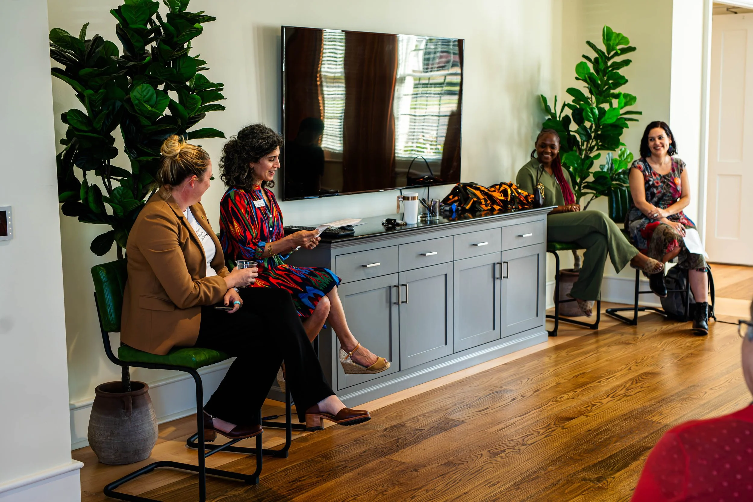 Four women sitting on chairs in a room with green plants, a large mirror, and a gray cabinet, engaged in conversation.