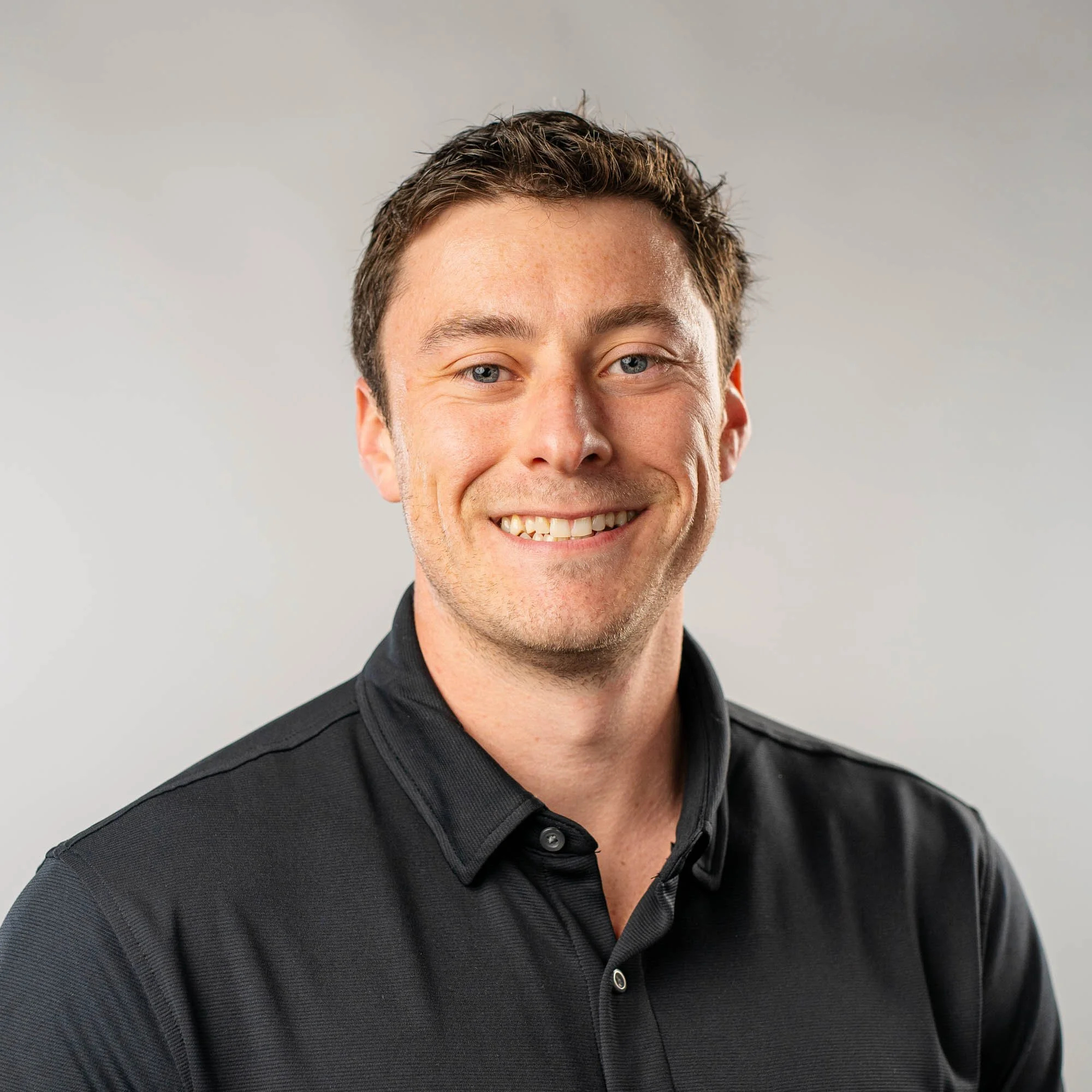 Close-up photo of a smiling young man with short brown hair, blue eyes, and fair skin, wearing a black collared shirt.