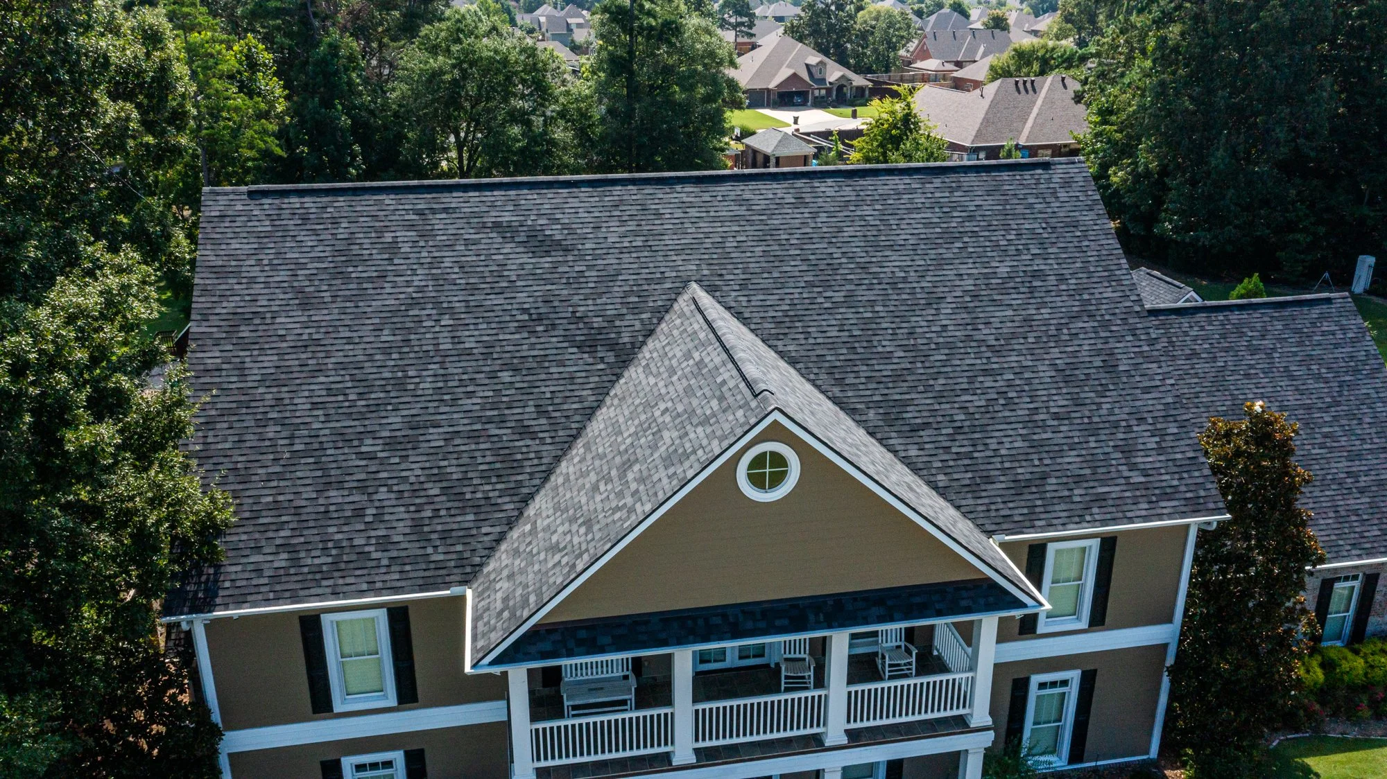 An aerial view of a house with a shingled roof and a balcony, surrounded by trees and neighboring houses.