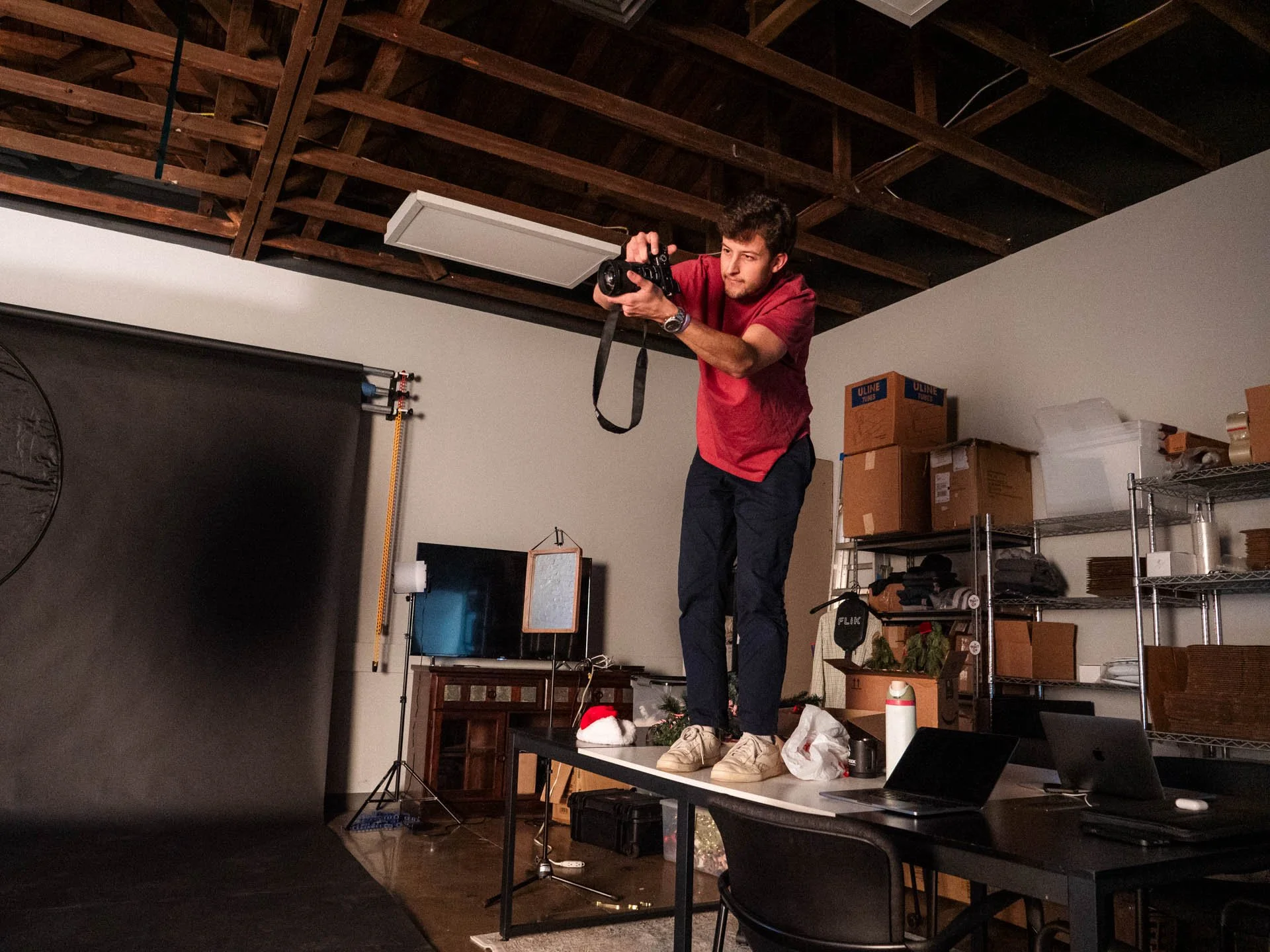 A man wearing a red shirt and beige shoes stands on a table, taking a photo with a camera. The scene appears to be a studio or workspace with some shelves, boxes, and equipment in the background.