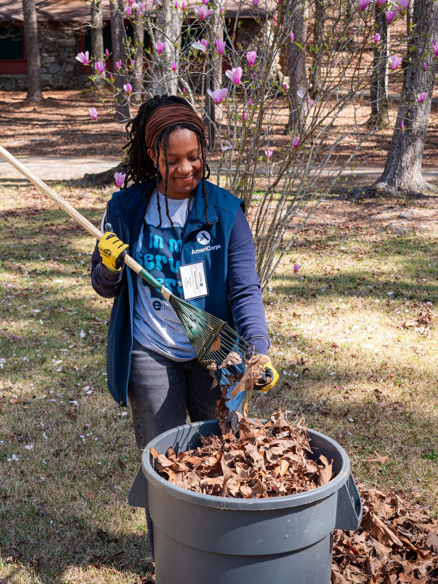A woman with dreadlocks, wearing a brown headband, blue vest, and gloves, is collecting fallen leaves into a large gray trash can outdoors surrounded by trees and blooming pink magnolia flowers.