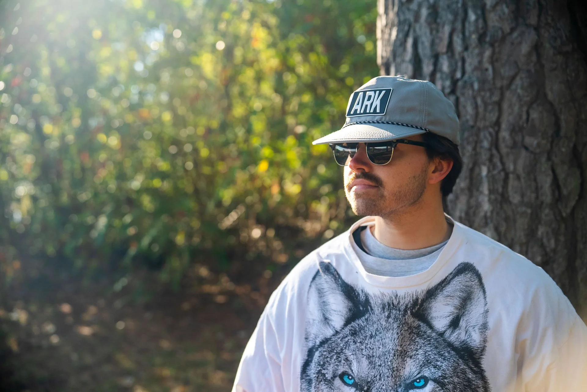 A man wearing sunglasses and a gray cap with the word 'ARK' on it, leaning against a tree outdoors in a forest setting, with sunlight filtering through the leaves.