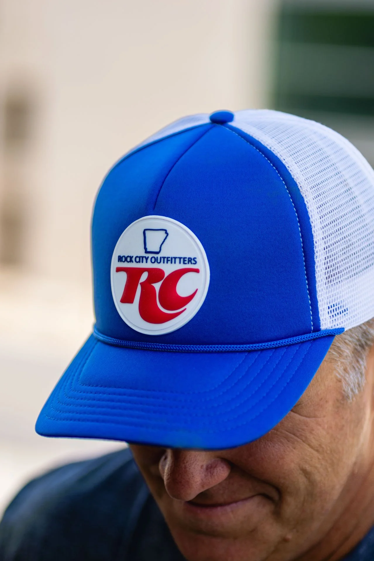 A person wearing a blue and white trucker-style cap with the Rock City Outfitters logo, partially covering their face, with visible gray hair and skin fold on their cheek.