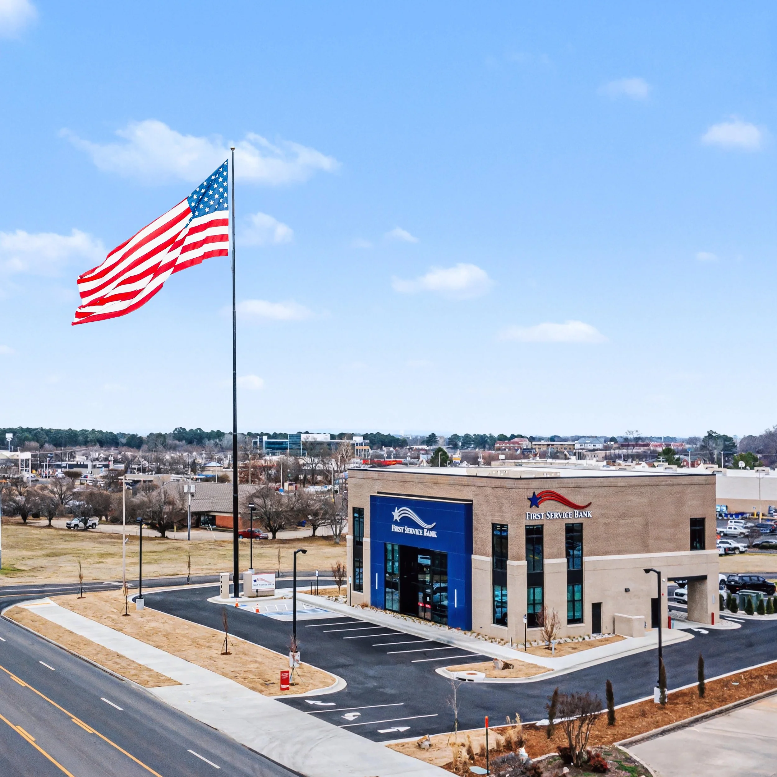 A Flag of the United States of America flying on a flagpole in a parking lot with new landscaping, a First Service Bank building, and a cityscape with residential and commercial buildings in the background under a partly cloudy sky.