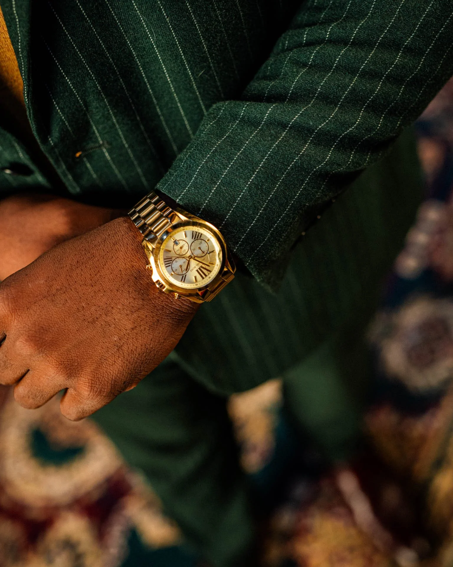 Close-up of a person's wrist wearing a gold watch with a white face and Roman numerals, against a background of a dark green pinstripe suit jacket.