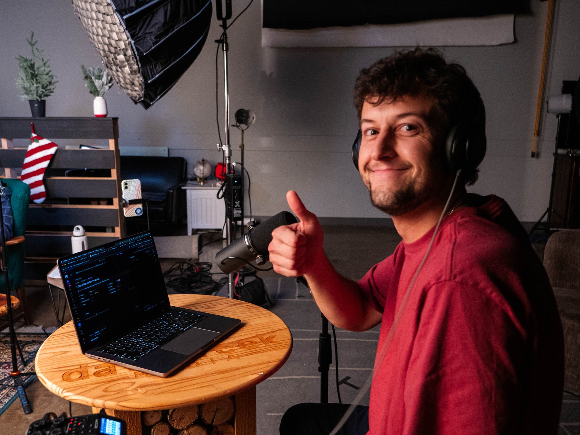 A man with curly hair and a beard wearing headphones, sitting at a round wooden table with a laptop, giving a thumbs up and smiling in a room with photography equipment and holiday decorations.