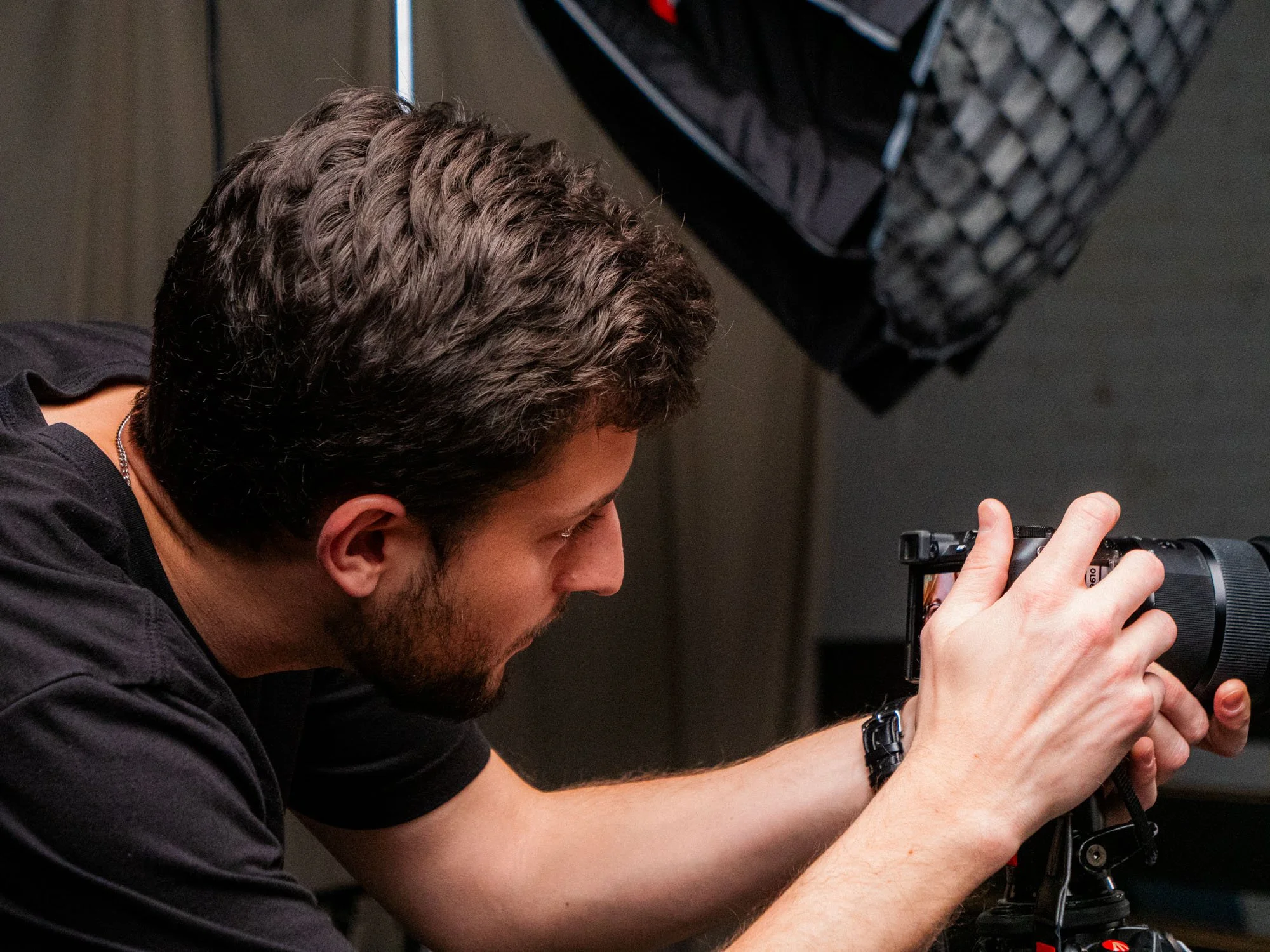 A young man with brown hair, a beard, wearing a watch, focused while using a camera on a tripod.