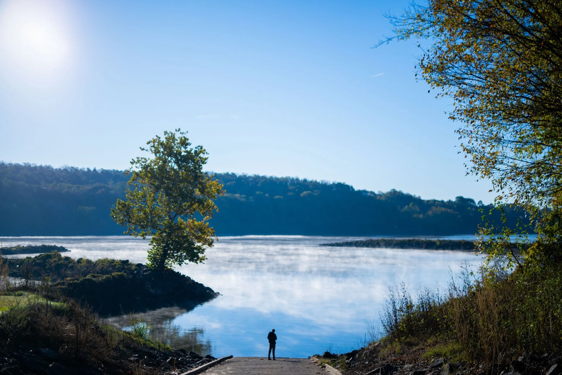 A person standing at the edge of a river, surrounded by trees with autumn leaves, with a backdrop of rolling hills and a clear blue sky.