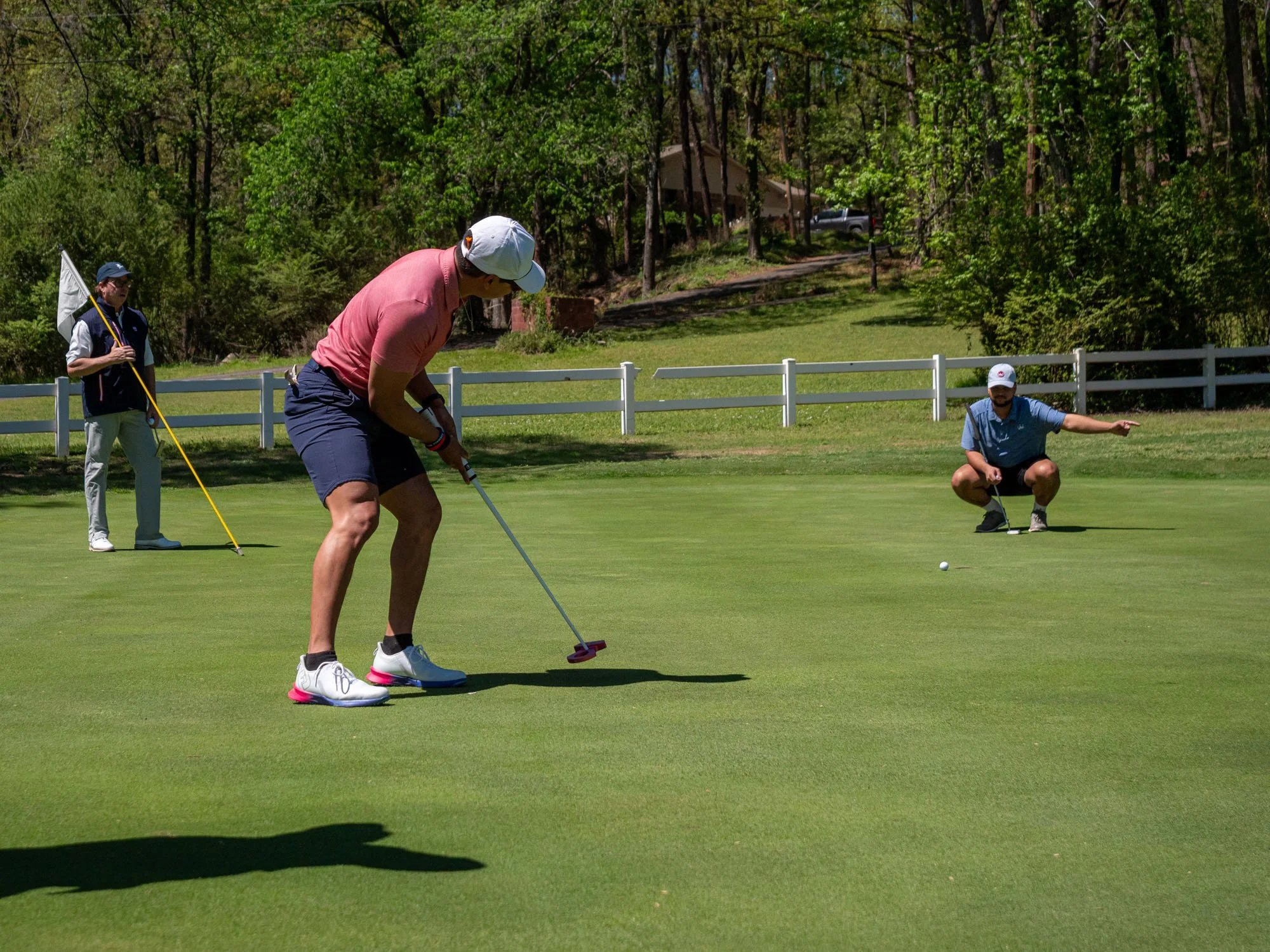 A woman in pink shirt and white cap putting golf ball on a putting green at a golf course, with two men in the background, one holding a flagstick and one crouching.