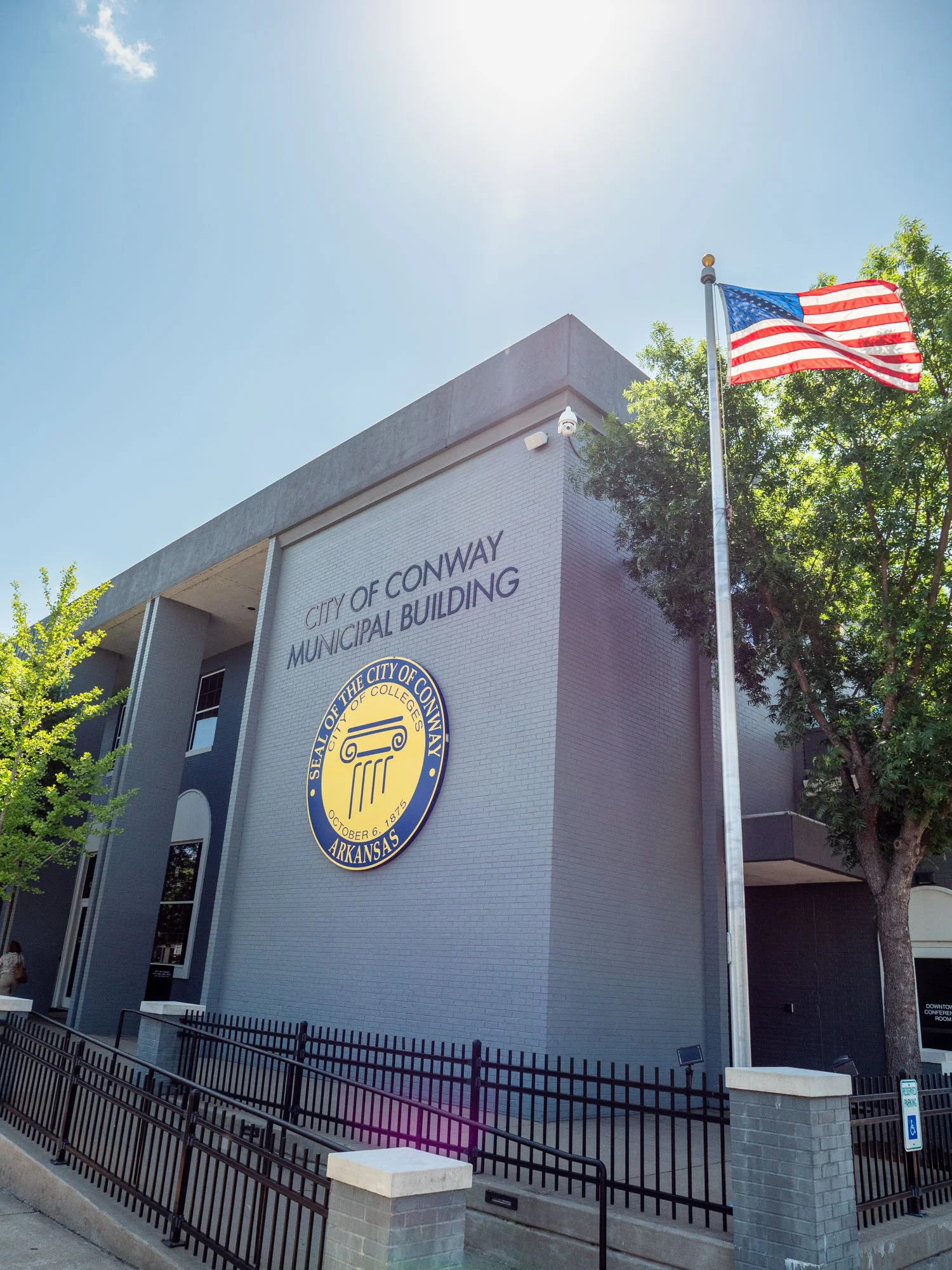 The City of Conway Municipal Building in Arkansas with an American flag on a flagpole, trees, and a clear blue sky.