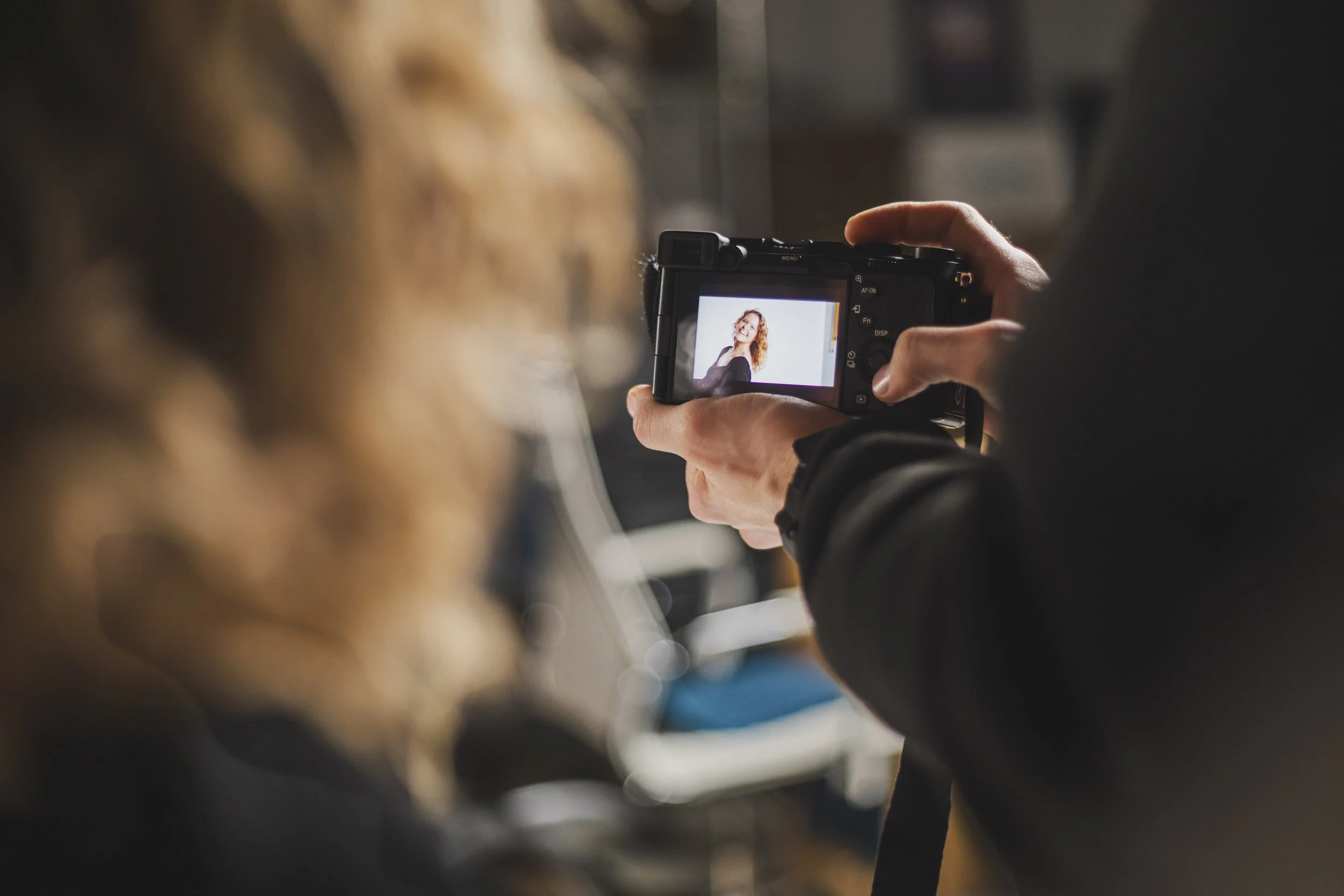 Person holding a camera, taking a picture of a woman who is smiling, with a blurred background.