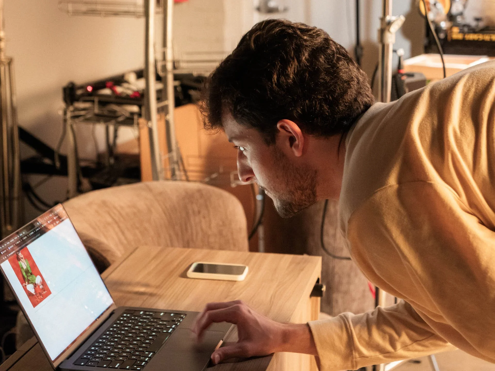 A man with dark, curly hair and a beard is leaning over a wooden table using a laptop. The laptop screen displays a colorful image related to Christmas. A smartphone is placed on the table next to the laptop. The background shows metal shelves and a 