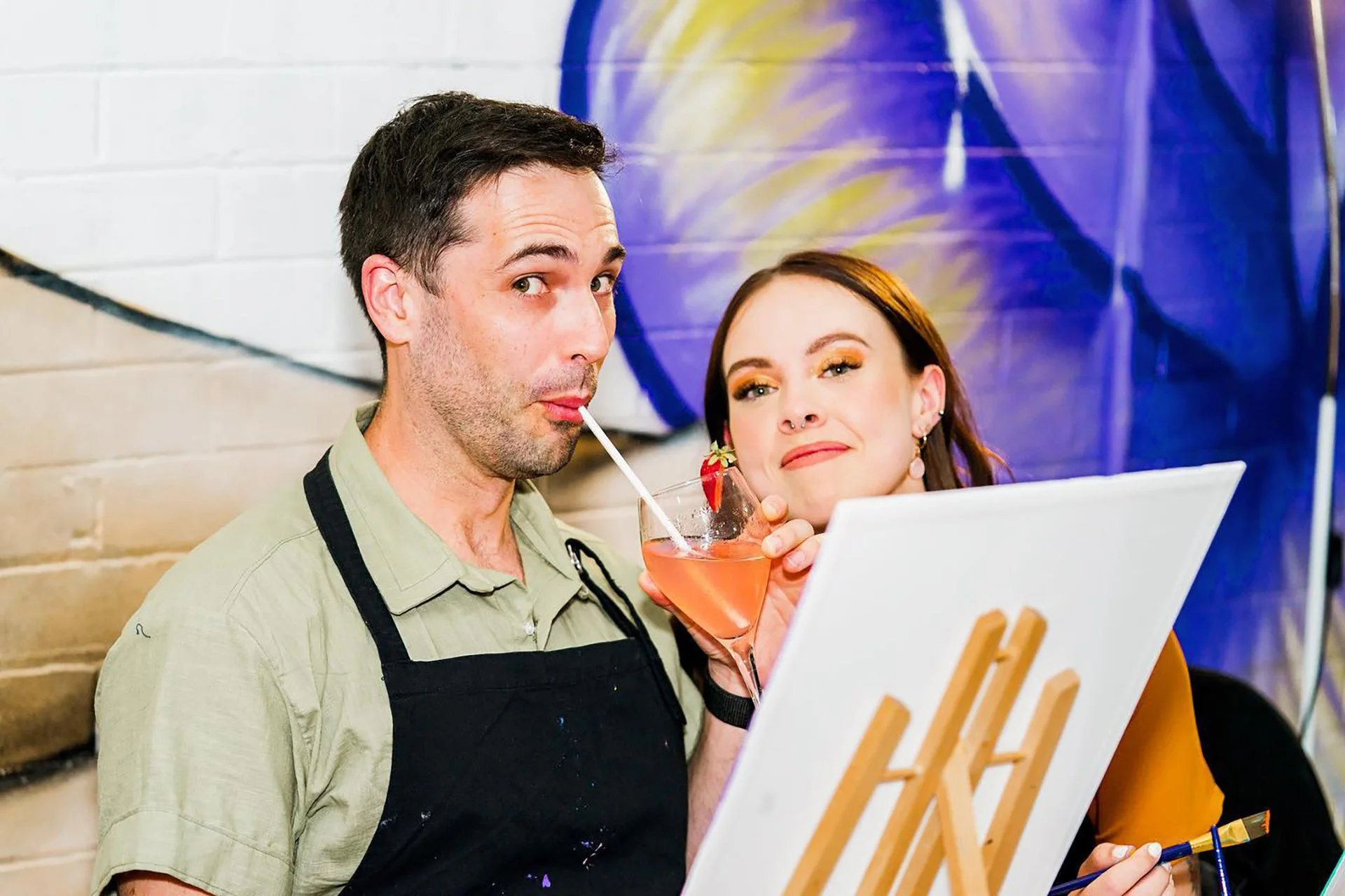 A man and a woman enjoying a drink, with the woman painting on an easel, at an art studio or gallery.